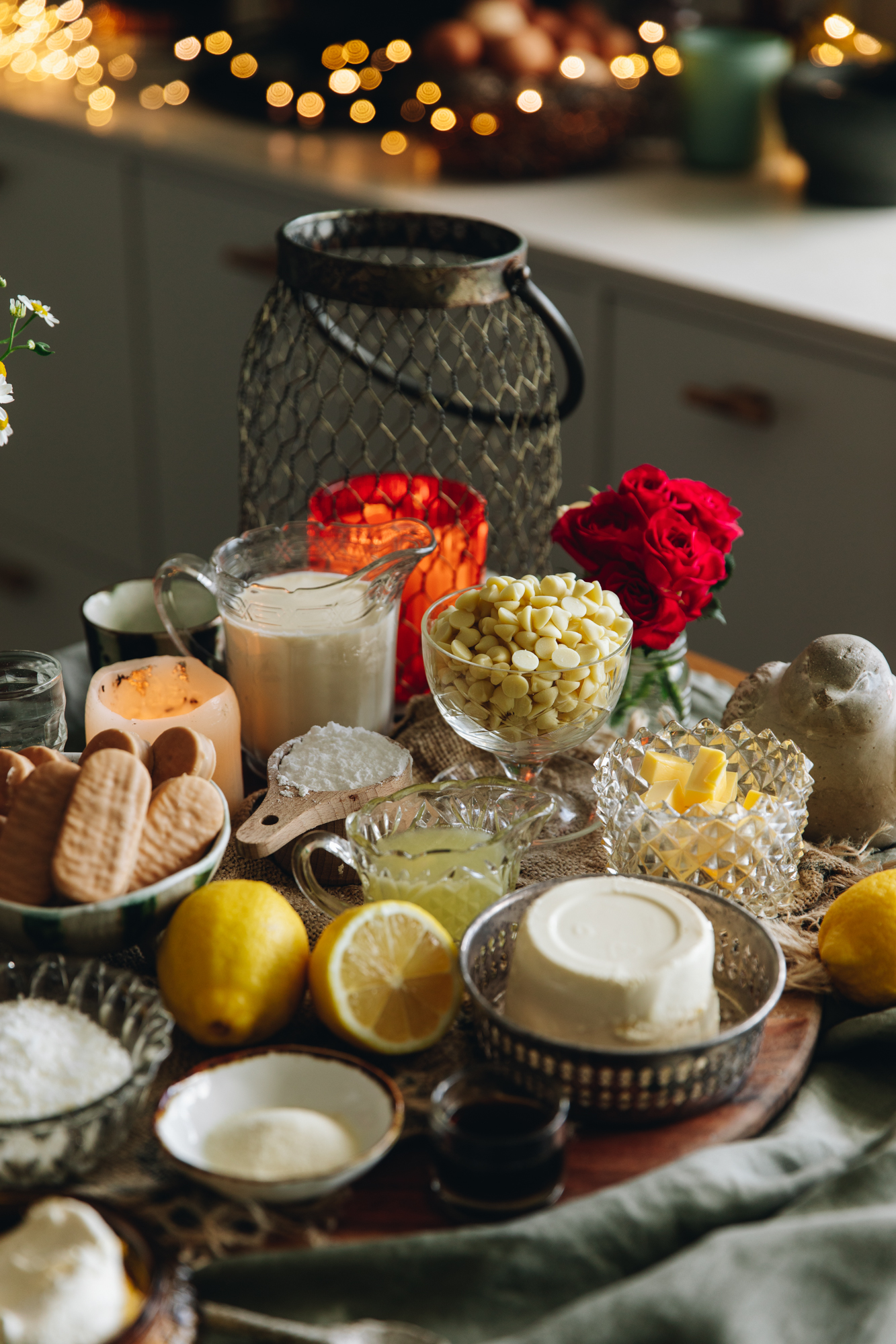 Cheesecake ingredients are displayed in vintage bowls and serve wear on top of a wooden board. Fairy lights are seen behind them.