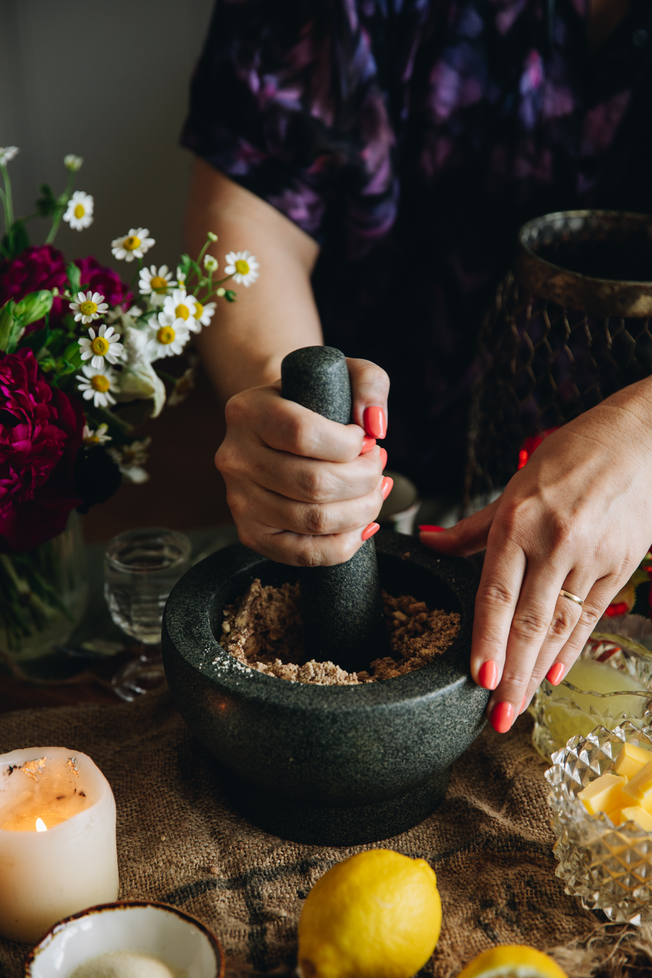 On a table is a mortar and pestle that is crushing white chocolate biscuits in to a crumb, a hand is holding the mortar and pestle.