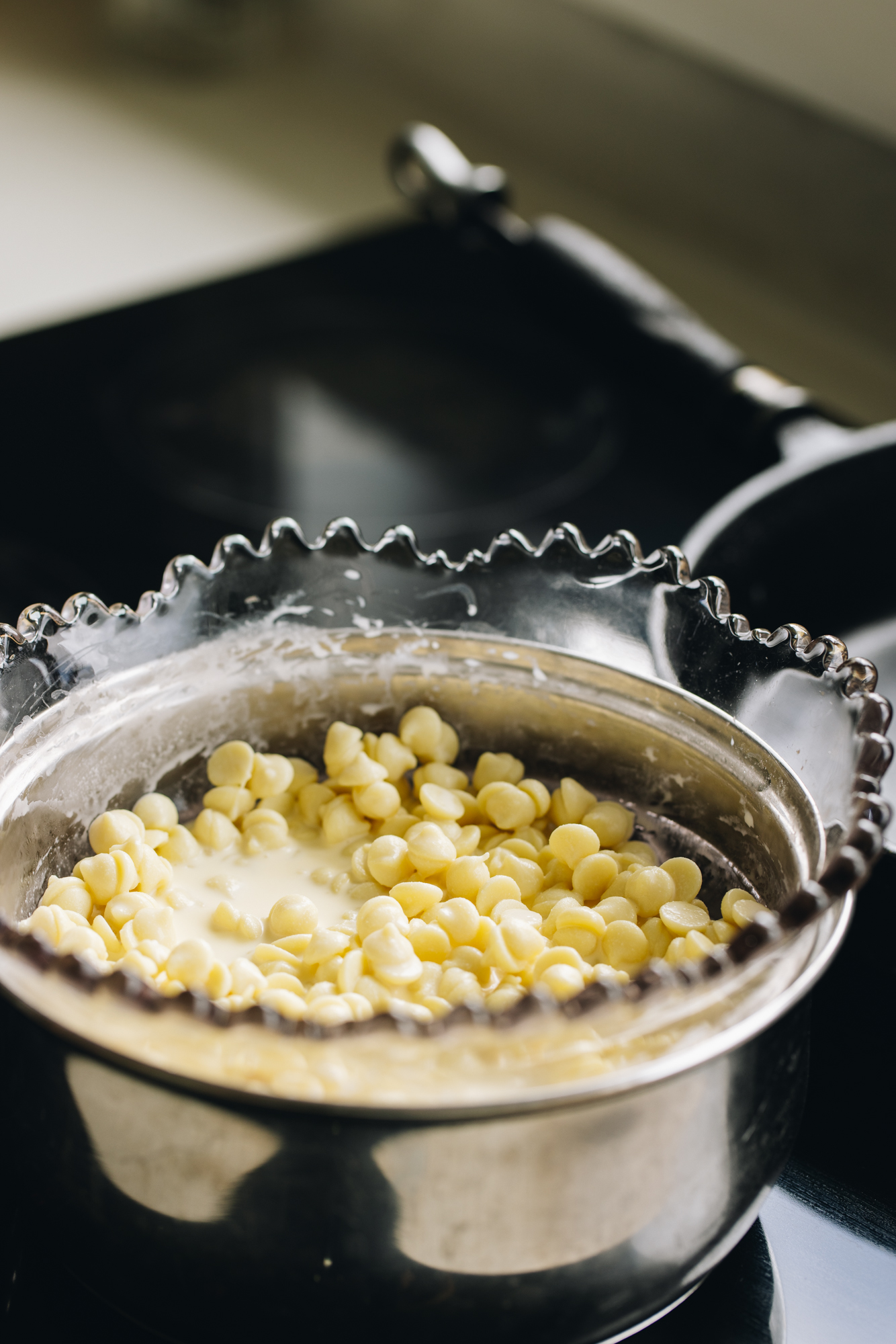 Chocolate drops and cream are set over a double boiler in a vintage jagged edge bowl, sitting in a pot on a black stove.