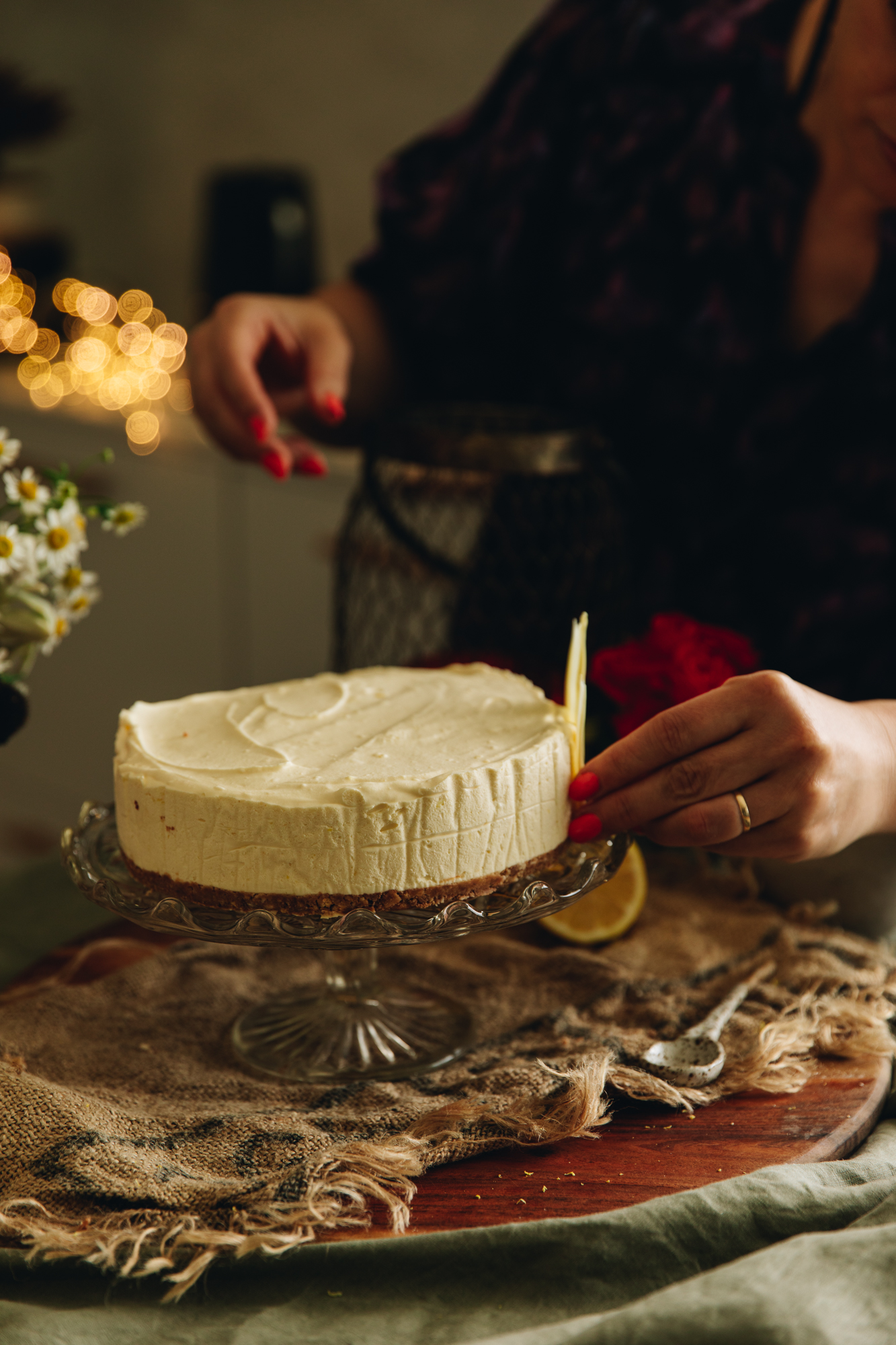 A hand is placing on a white chocolate petal decoration on the plain lemon and white chocolate cheesecake that is on a glass cake stand, on a wooden board and rustic cloth.