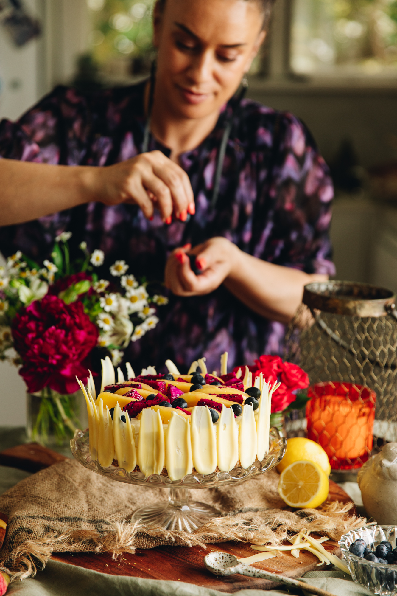 Naomi Toilalo is holding in the background of the lemon and white chocolate cheesecake that is on a glass cake stand, on a wooden board and rustic cloth. It has mango and dragon fruit arranged on top. Flowers are next to Naomi.