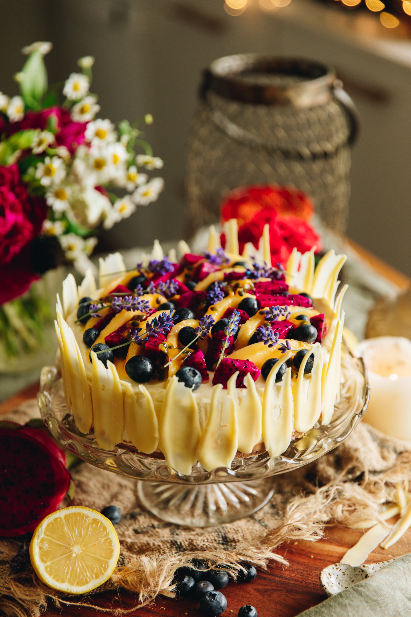 The lemon and white chocolate cheesecake is on a glass cake stand, on a wooden board and rustic cloth. It has mango and dragon fruit, blueberries and lavender flowers arranged on top. Flowers are next to the cheesecake.