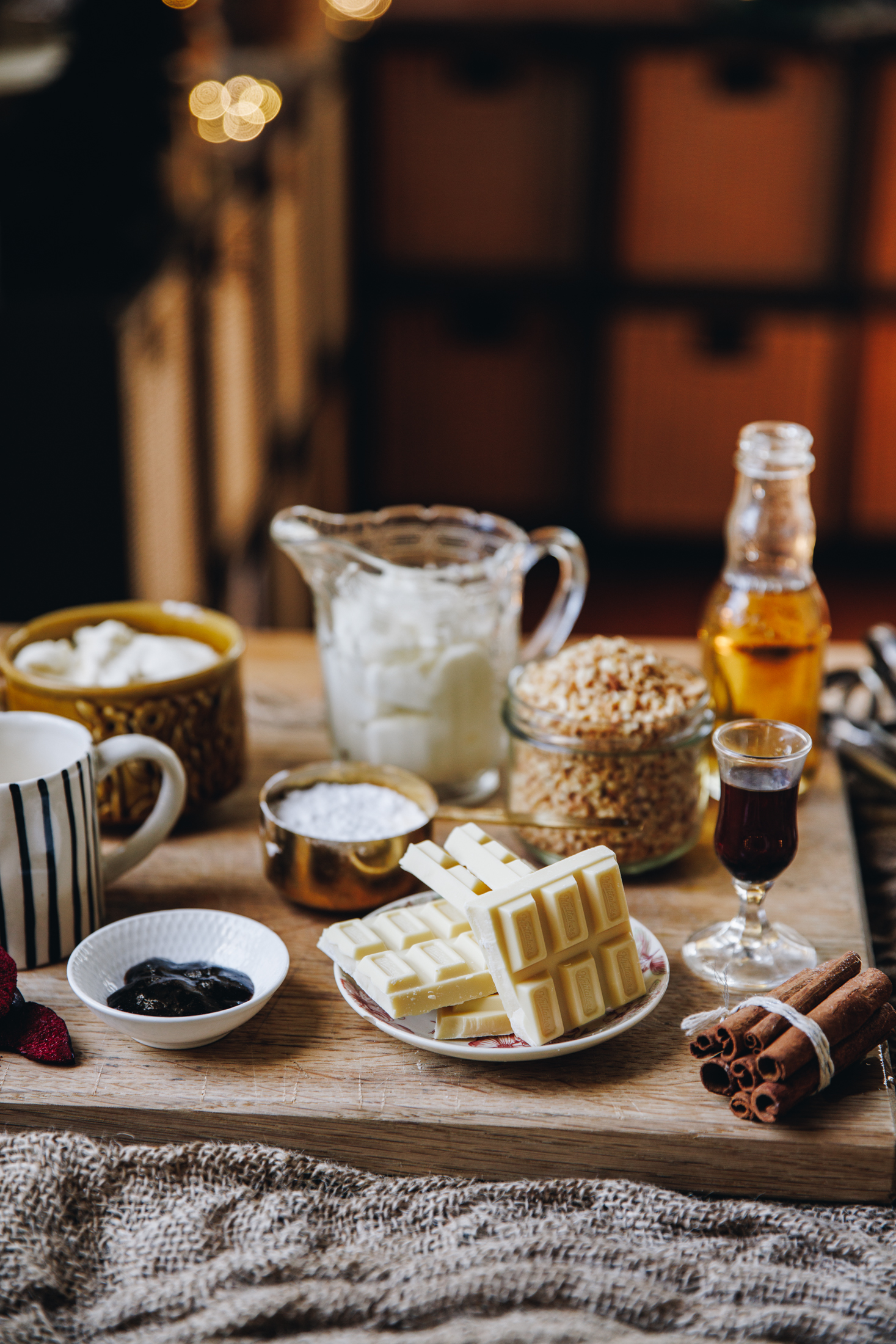 White chocolate ingredients are displayed in vintage bowls and serve wear on top of a wooden board. Fairy lights are seen behind them.