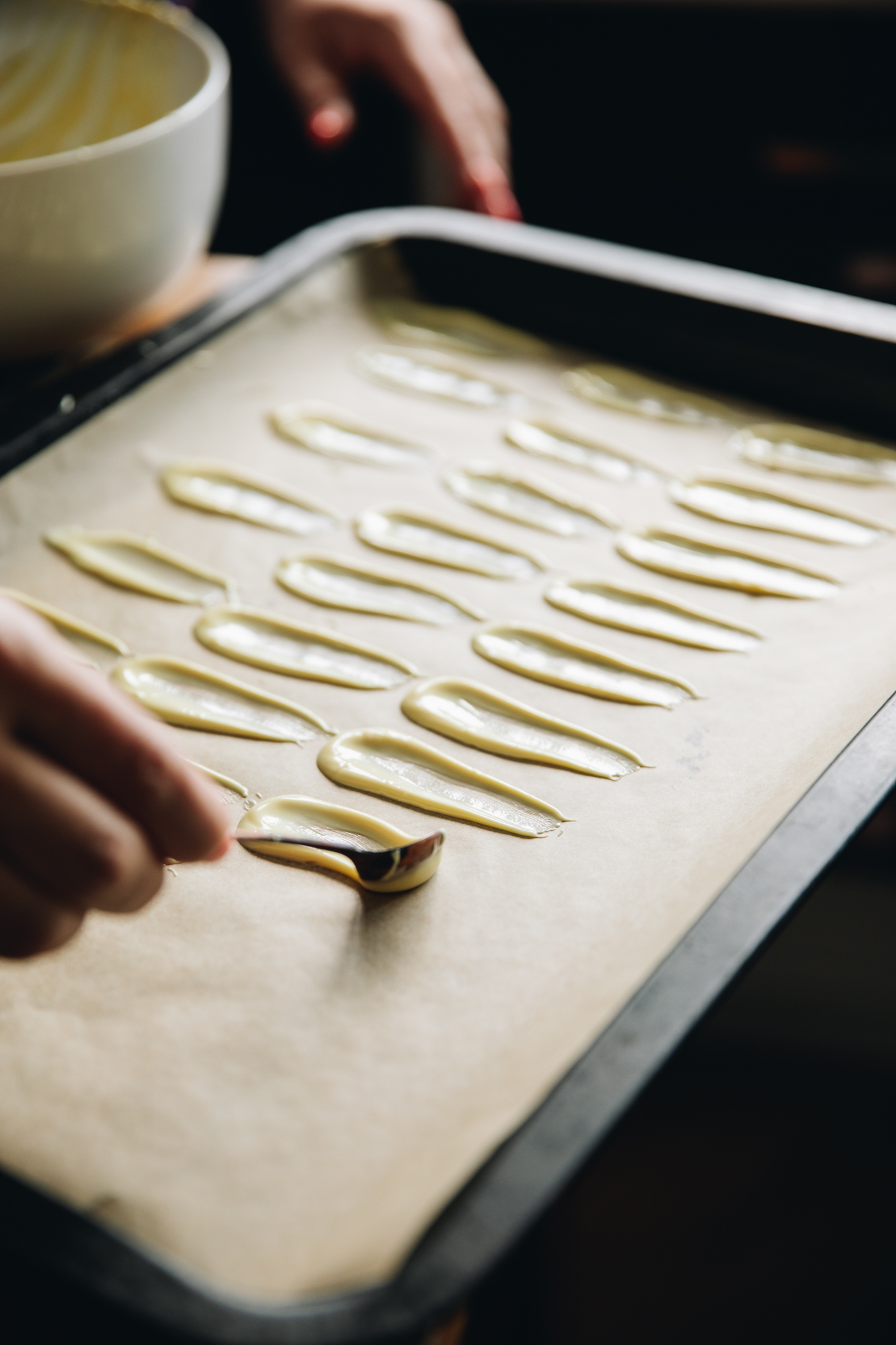 On a tray with brown baking paper white chocolate is being spread on in small petals shapes. The tray is full with many petals.