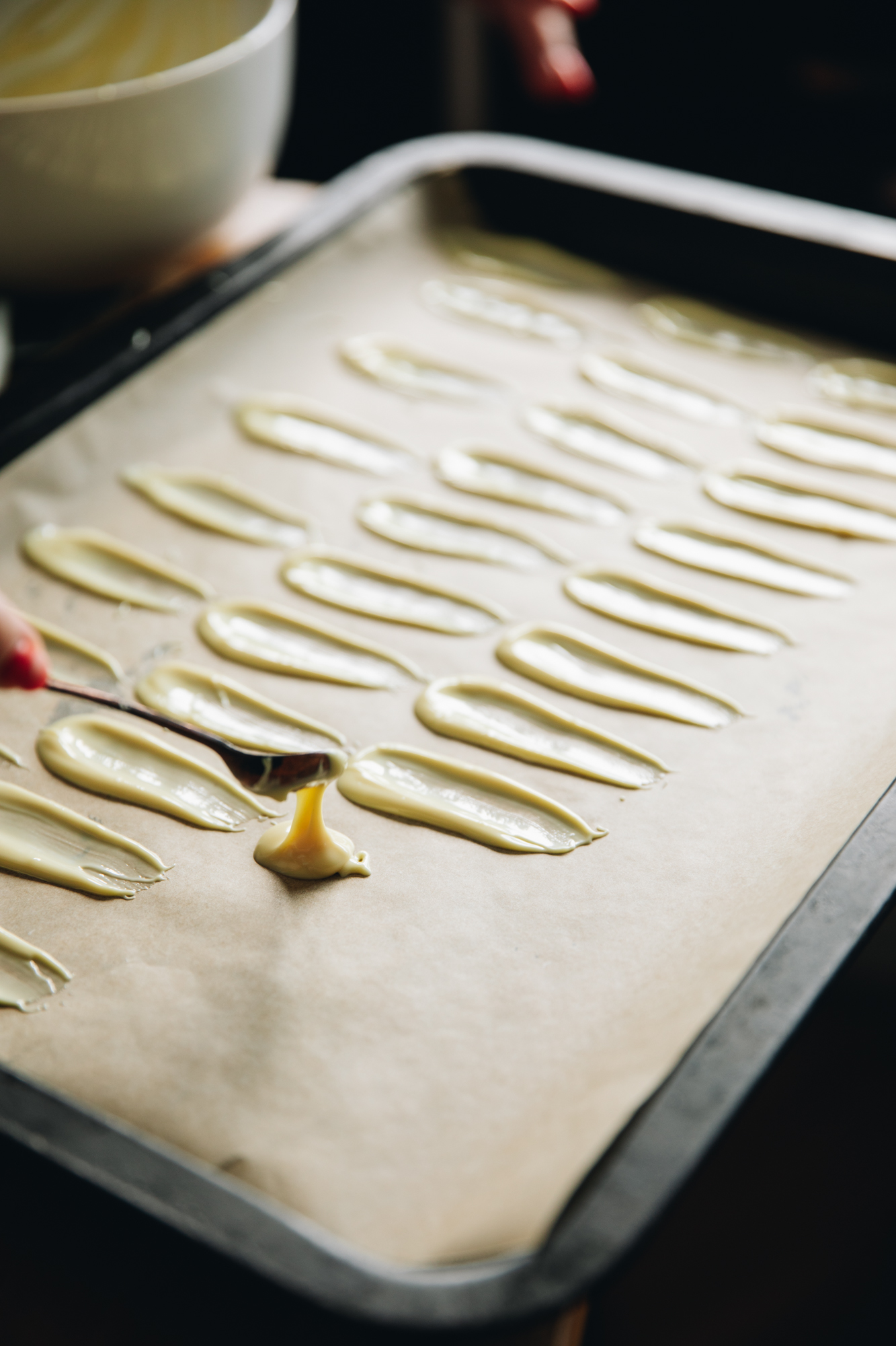 On a tray with brown baking paper white chocolate is being spread on in small petals shapes. The tray is full with many petals.