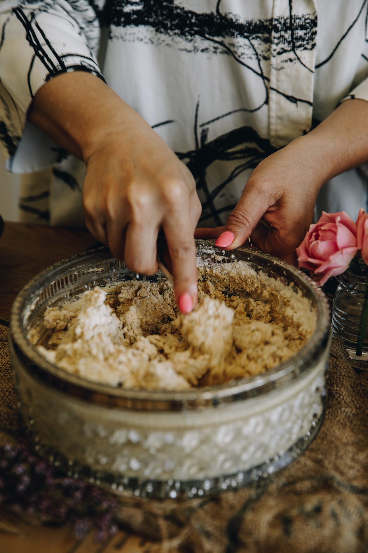 On a wooden table with natural fabric on it sits a glass vintage bowl with a silver trim.In it is biscuit mixture being stirred with a fork by Naomi.
