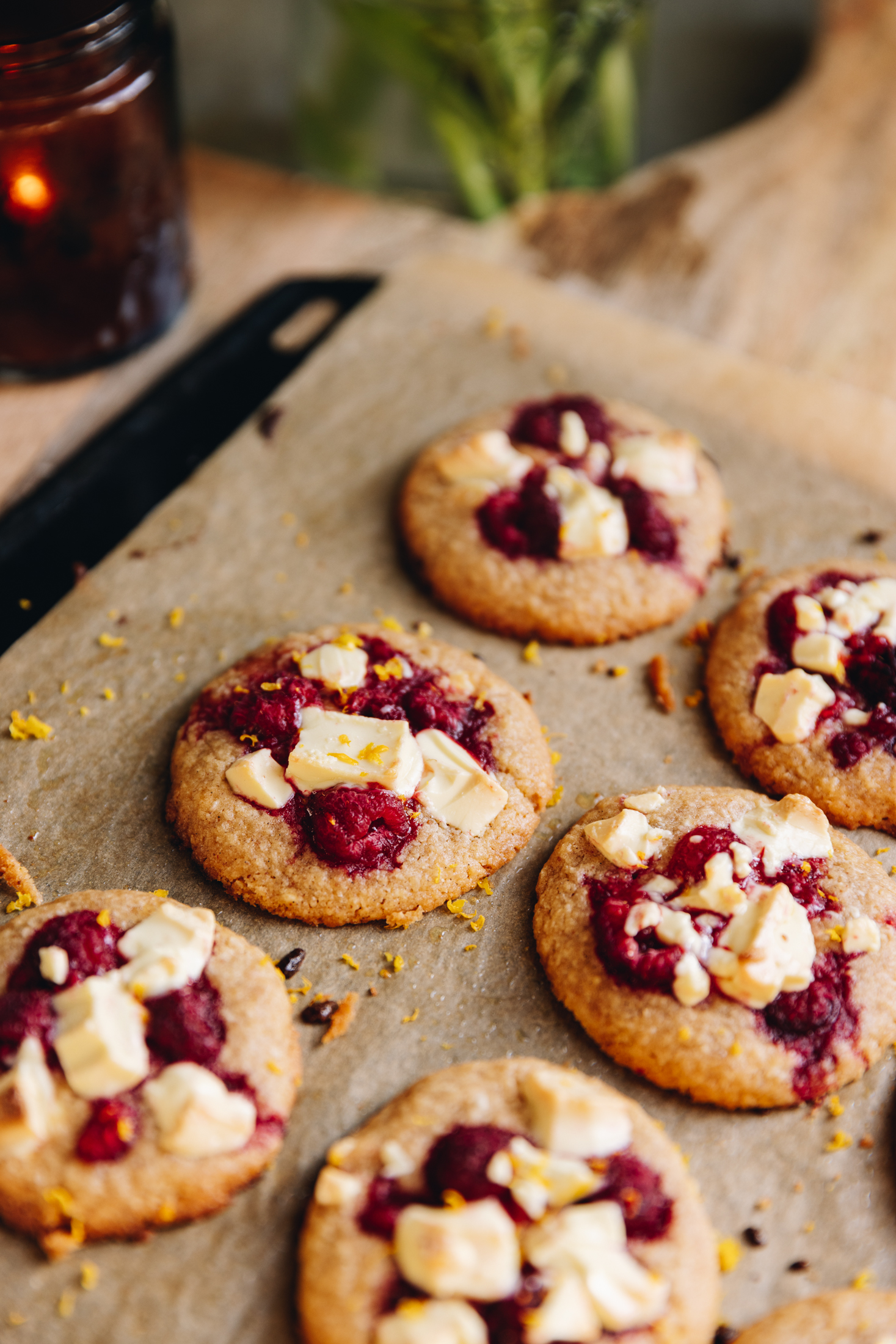 Freshly baked white chocolate raspberry biscuits are on a black tray lined with baking paper. An amber candle burns in the background. 