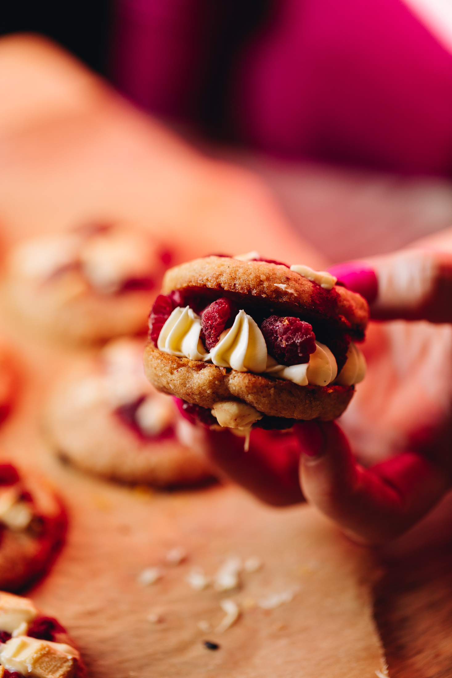 A close up shot reveals a lemon raspberry cookie sandwich which has two white chocolate raspberry cookies with pipes cream in the middle with pieces of fresh raspberry.