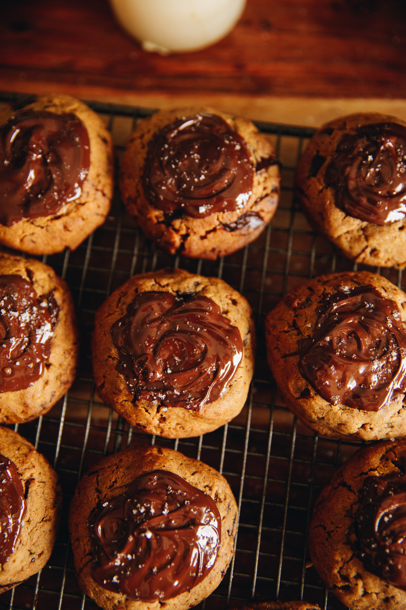 A flat lay shot shows freshly baked Dark Chocolate Molasses Cookies. They are on a wire cooling rack, on top of a wooden table. They have been decorated with a smear of dark chocolate and a sprinkle of flaky sea salt.