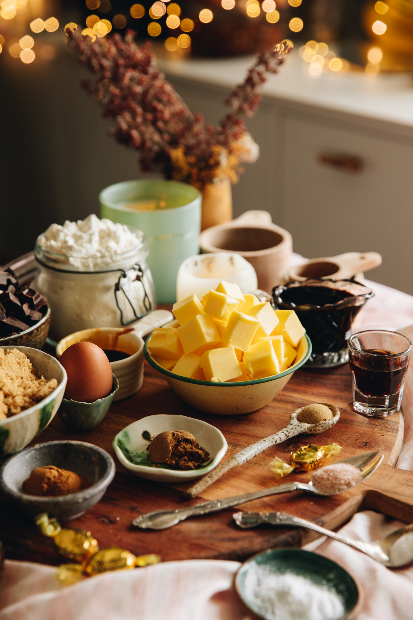 A round wooden board sits on a table lined with a soft pink tablecloth. On the board is cookie ingredients placed in vintage bowls, spoons and jars. In view is flour, butter, eggs, spices, brown sugar, vanilla and molasses. In the background is flowers and a blue candle burning. Fairy lights are in the background.