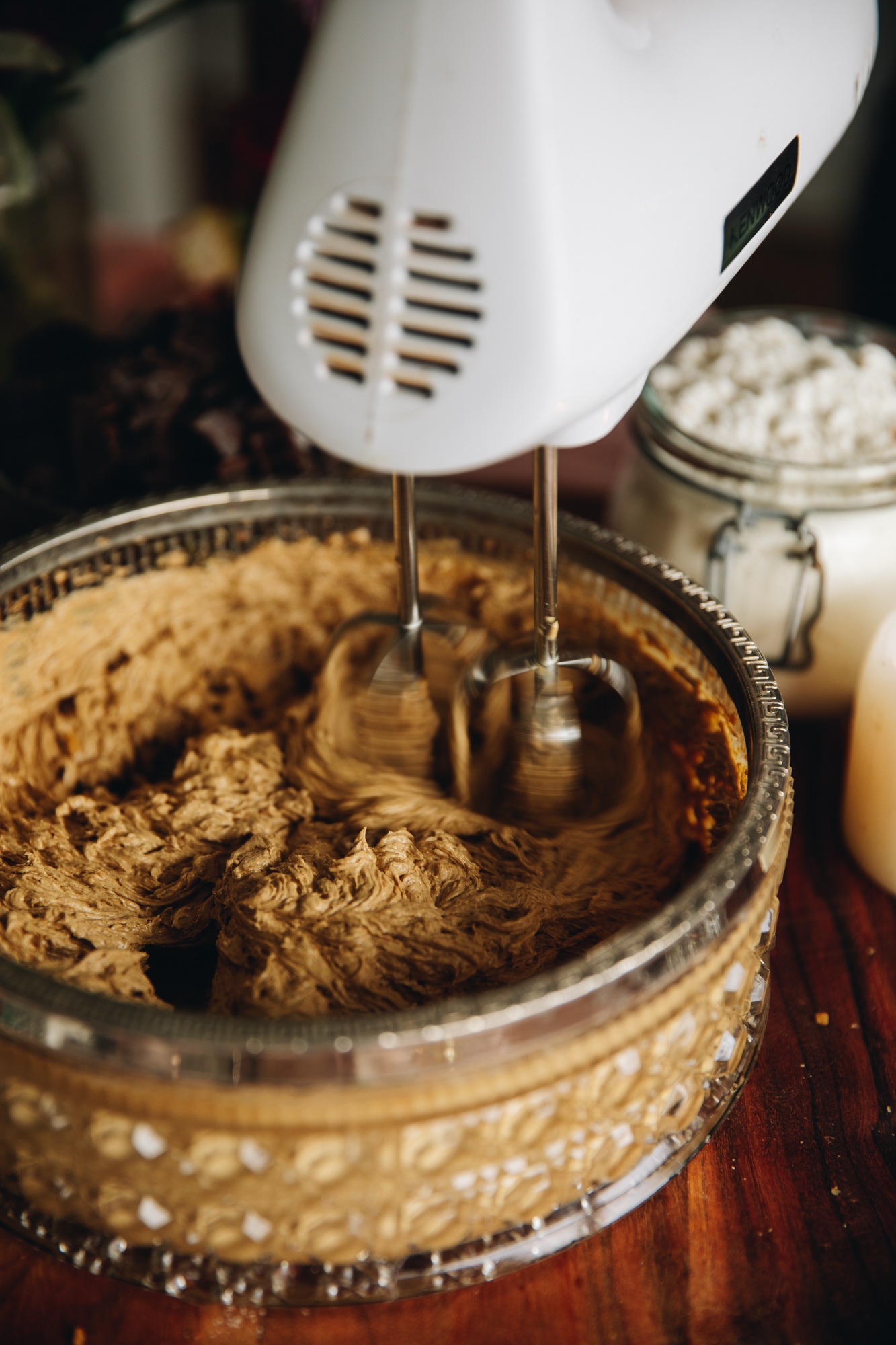 A vintage bowl with a silver trim sits on top of a striped wooden board. In the bowl is butter, molasses, spices and sugar is being whipped by a white hand mixer. A jar of flour is in the background with a candle next to it.