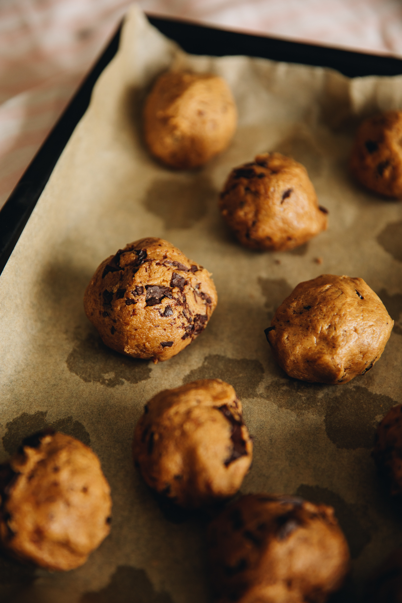 A black tray sits on top of a soft pink tablecloth, on the cloths sits a black baking tray lined with baking paper with unbaked dark chocolate molasses cookies balls.