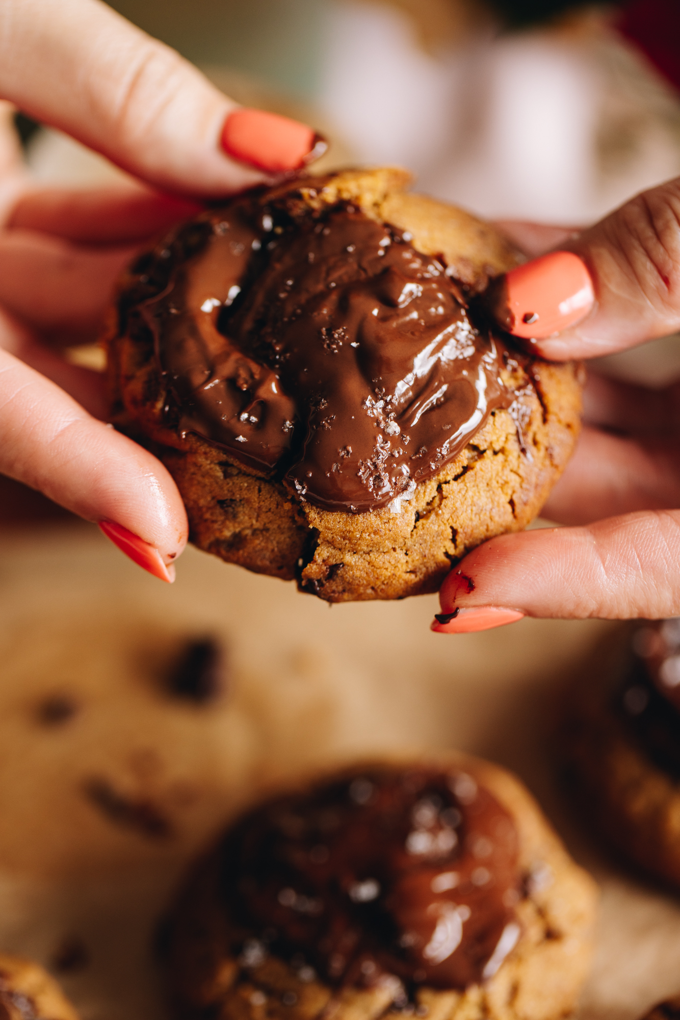 An extreme close up shot of freshly baked dark chocolate molasses cookies is held up and is being broken by Naomi's hands to reveal the gooey chocolate that is topping the biscuit.