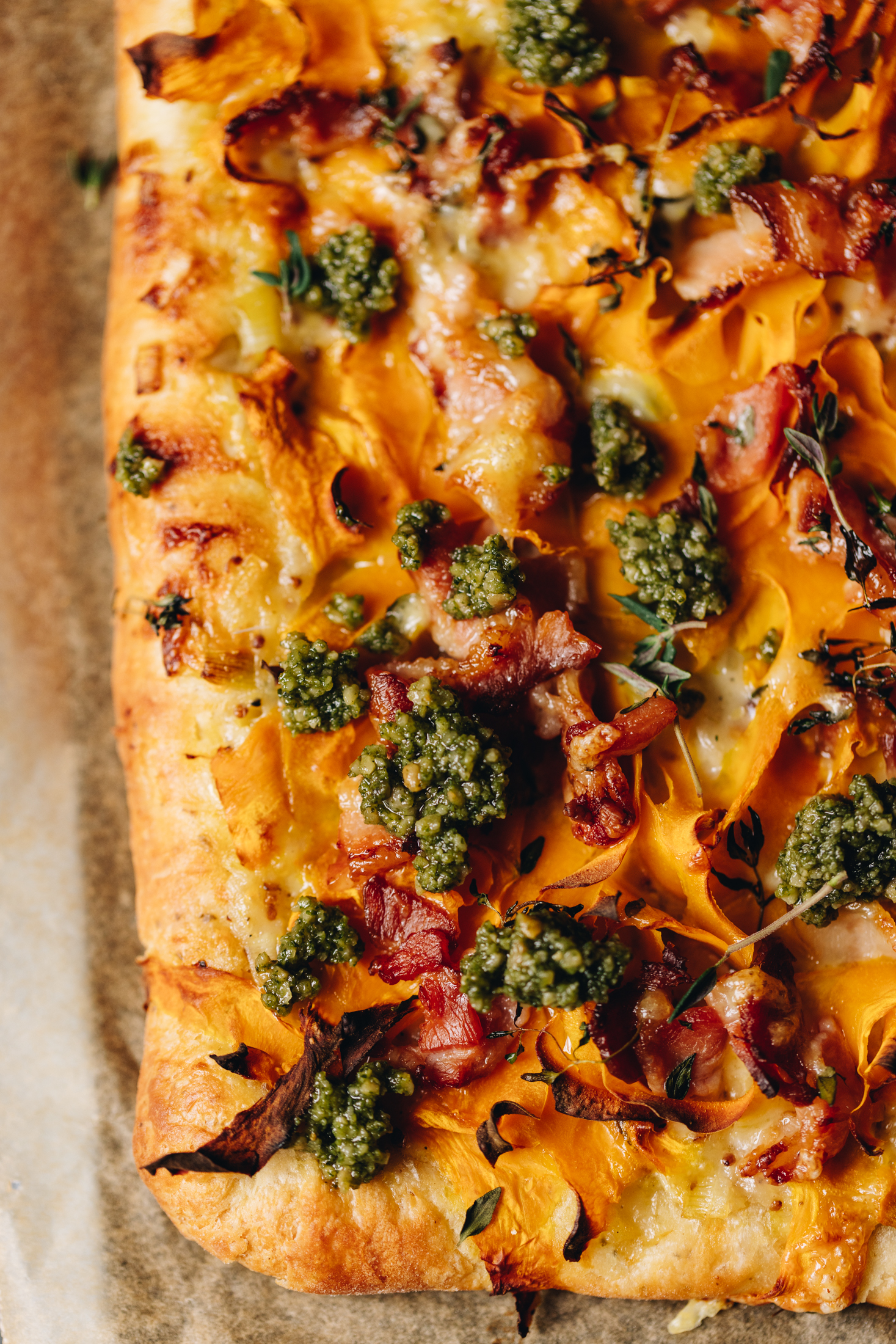 A flat lay shows a baked creamy pumpkin tart that has crispy pastry underneath it. On top of the tart is dollops of pesto, melted cheese, pumpkin ribbons and a little bit of bacon.