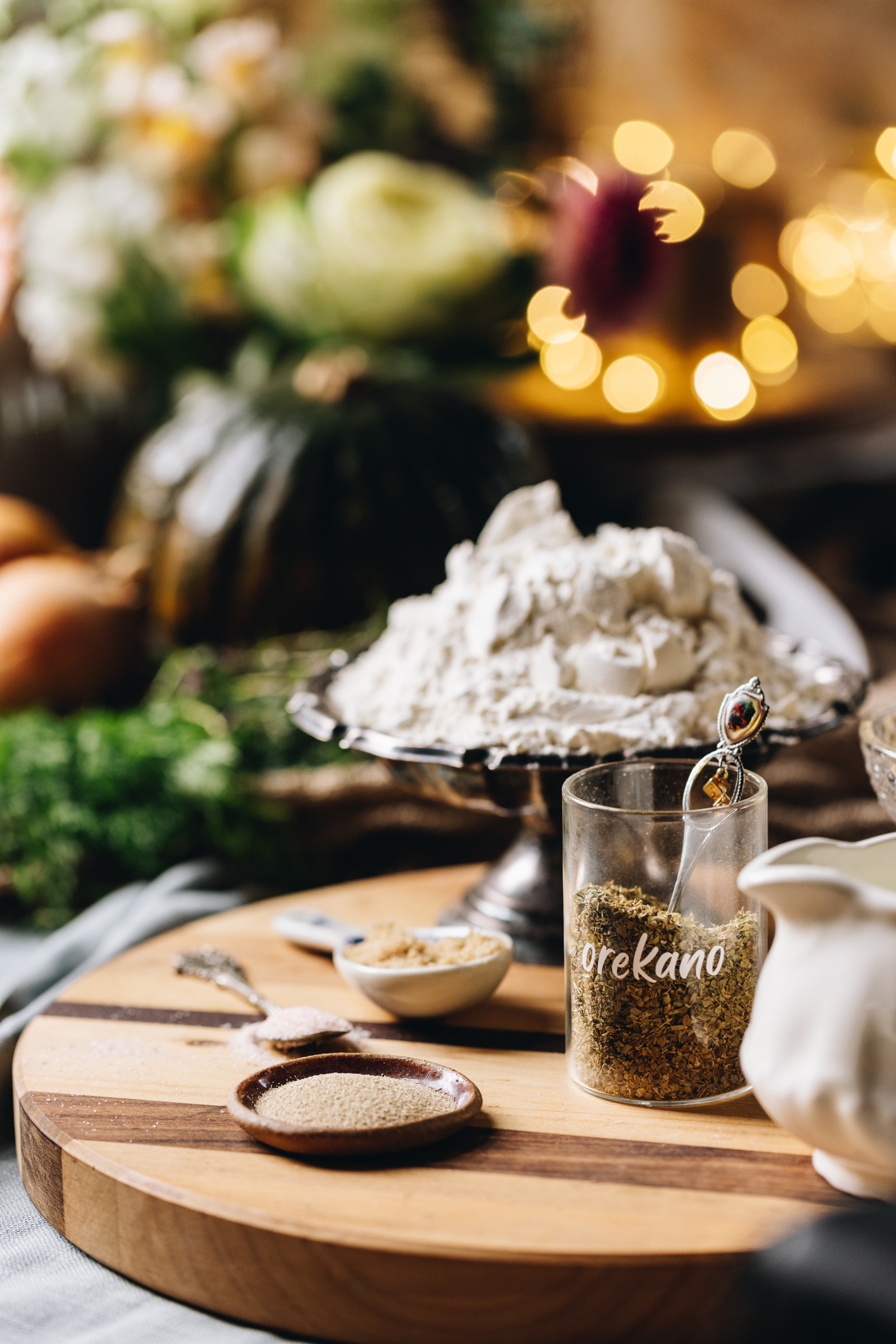 A wooden striped board is on a grey tablecloth. On the board is a jar of oregano, yes, a white jug, a ceramic spoon and a silver bowl with flour in it. In the background is onions, a pumpkin and flowers all blurred out with the glow of fairy lights in the background. 