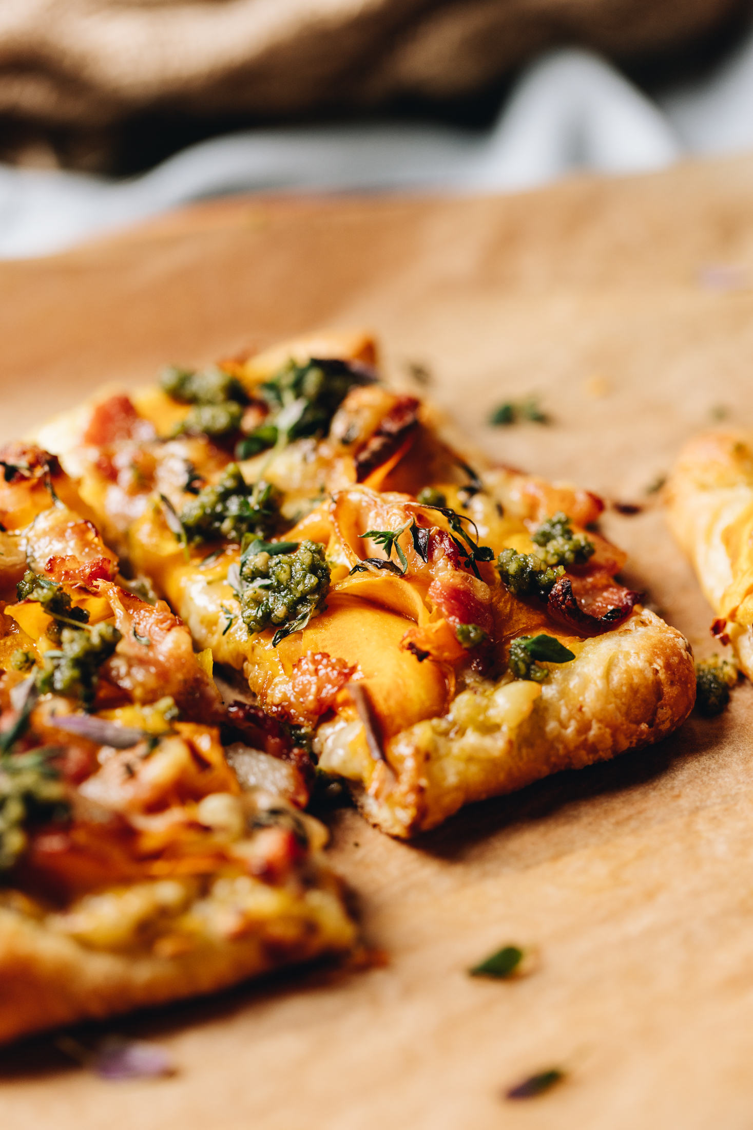 Crunchy yeasted savoury pastry has been baked in to mini pumpkin tarts. On the pastry is slivers of pumpkin, bacon, thyme, cheese and green pesto. A mini tart has been cut in half and the pastry is golden.