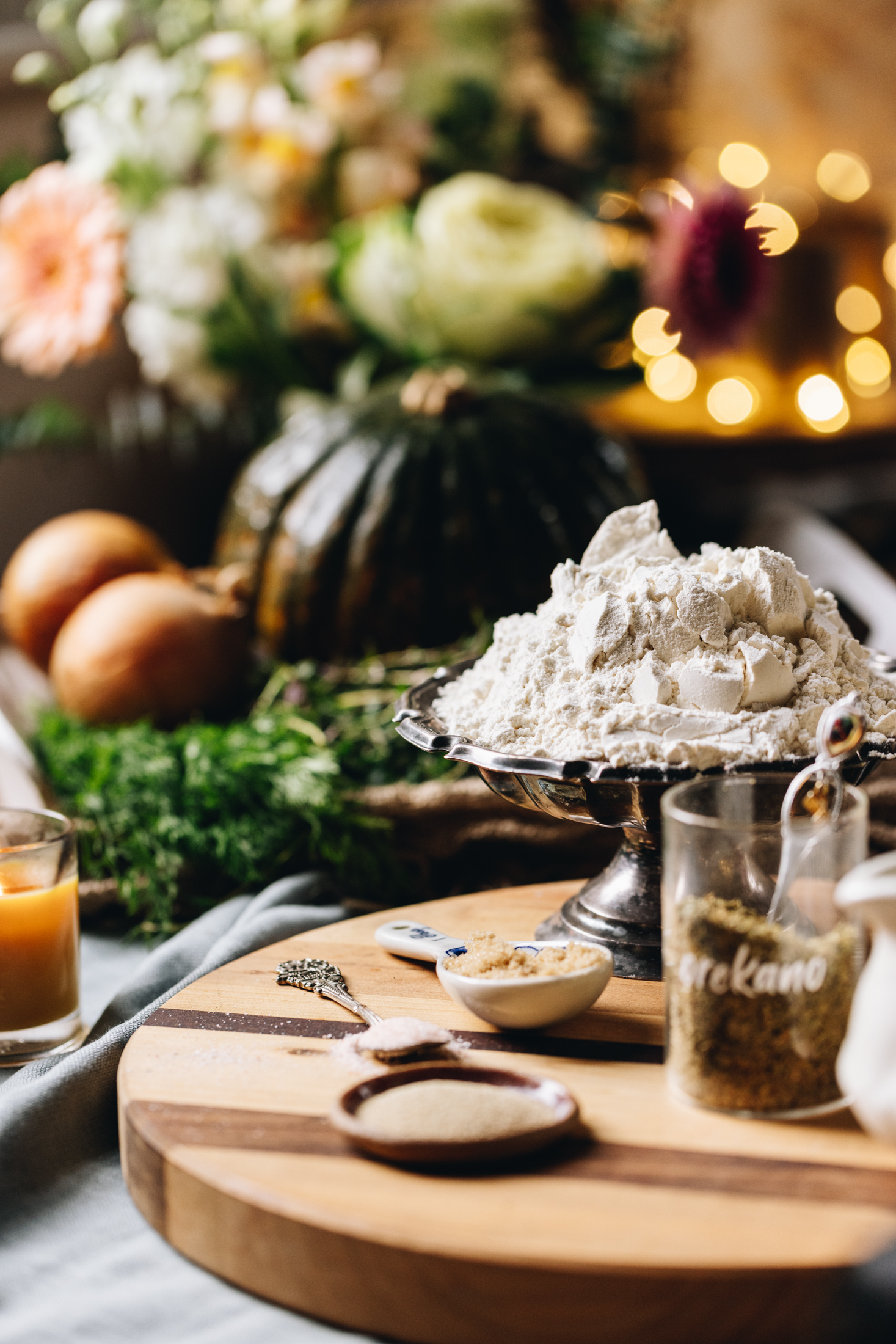 A wooden striped board is on a grey tablecloth. On the board is a jar of oregano, yes, a white jug, a ceramic spoon and a silver bowl with flour in it. In the background is onions, a pumpkin and flowers all blurred out with the glow of fairy lights in the background.