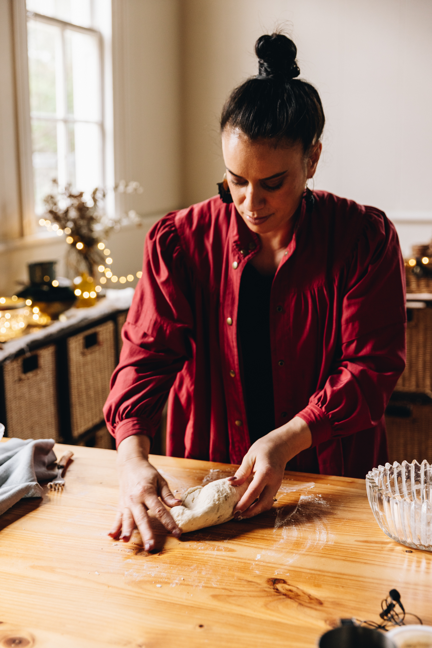 Naomi Toilalo is kneading savoury pastry on a wooden table. Behdin her is golden fairy lights and a tea towel to her side. I glass vintage bowl is on the other side of her.