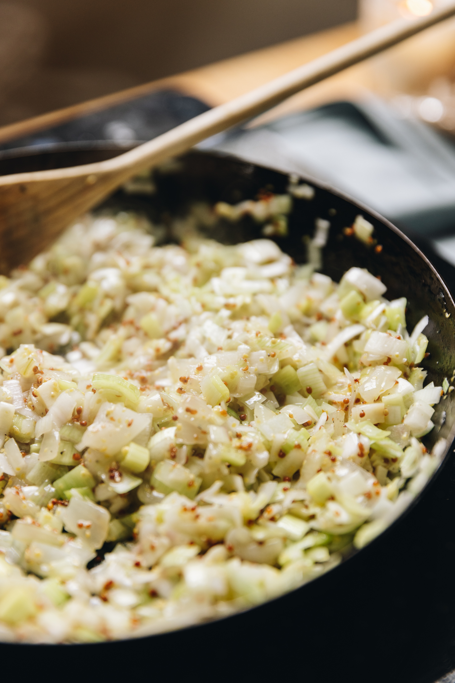 A black cast iron pan sits on a black portable stove, on a wooden table. In the pan is diced onions and wholegrain mustard that is cooking. A wooden spoon is stirring it.