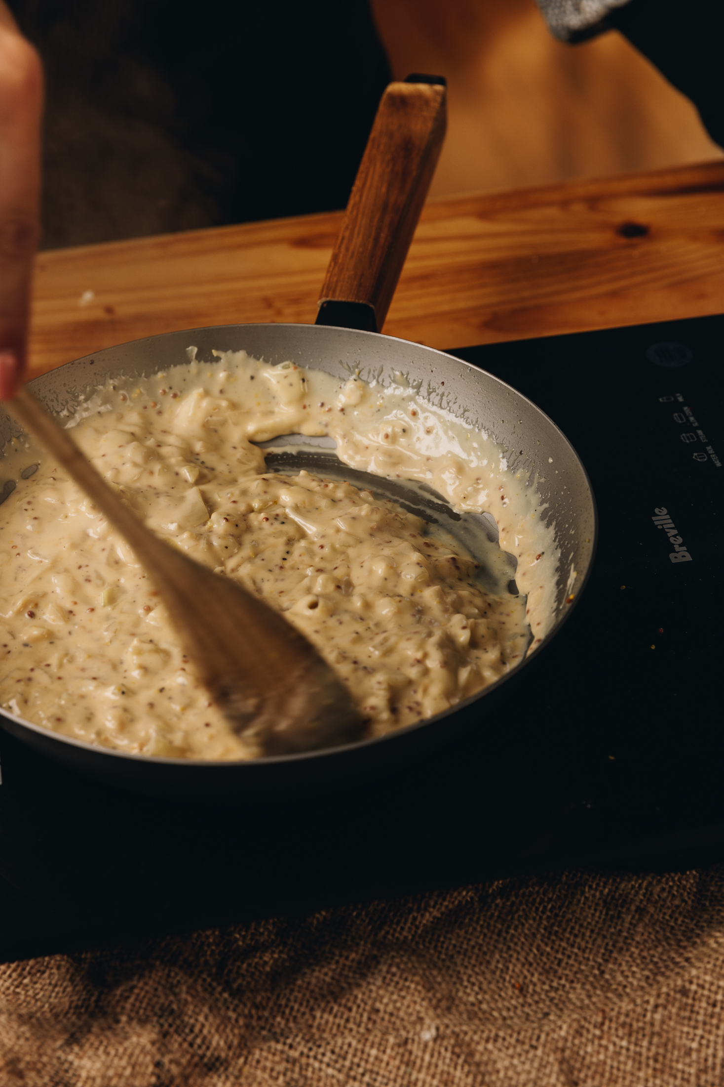A black cast iron pan sits on a black portable stove, on a wooden table. In the pan is a creamy onion and cheese mixture that is being stirred with a wooden spoon.
