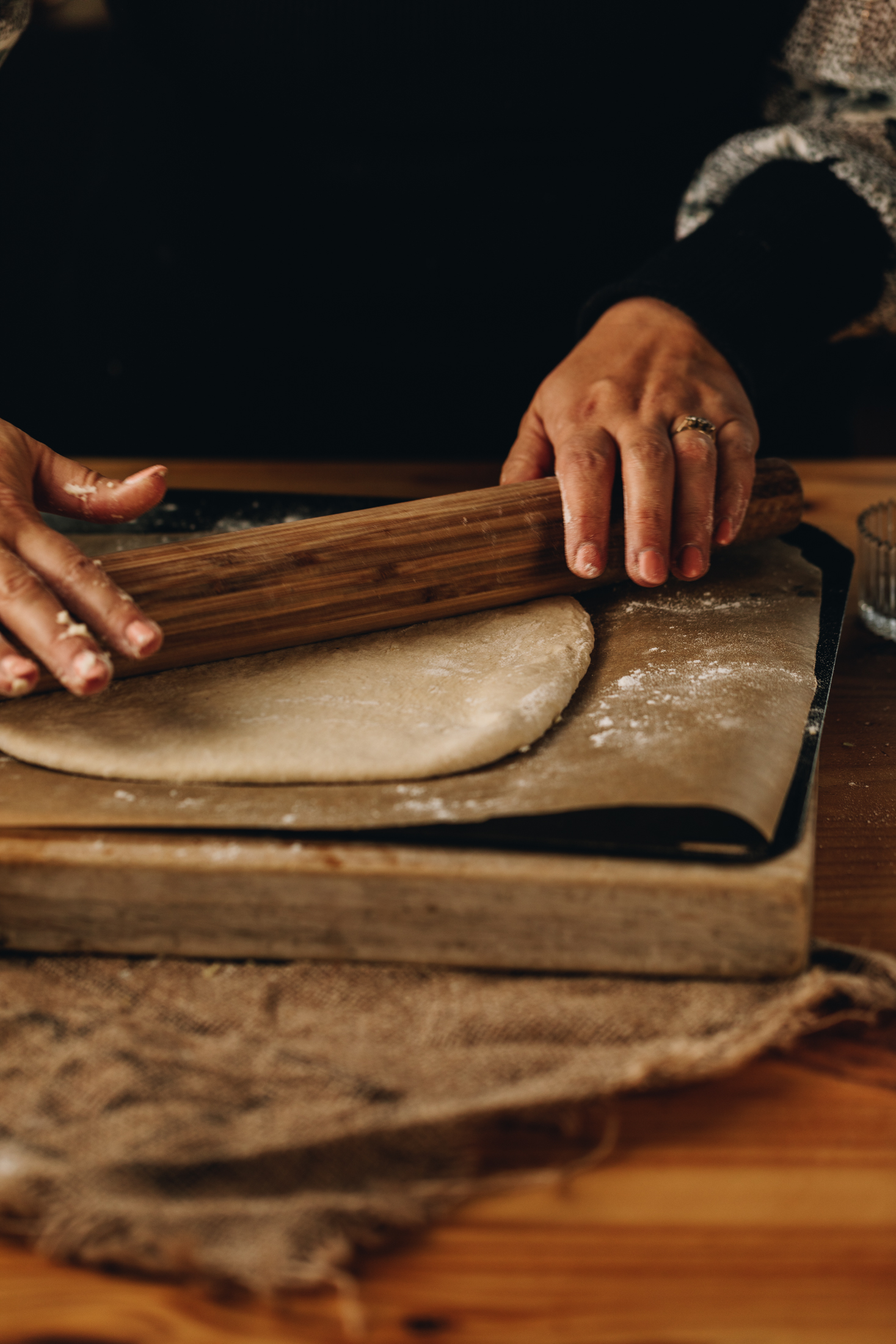 A wooden board is on a natural piece of fabric, on a wooden table. On the board is a black tray lined with brown baking paper and Naomi is rolling out a pastry on to it.