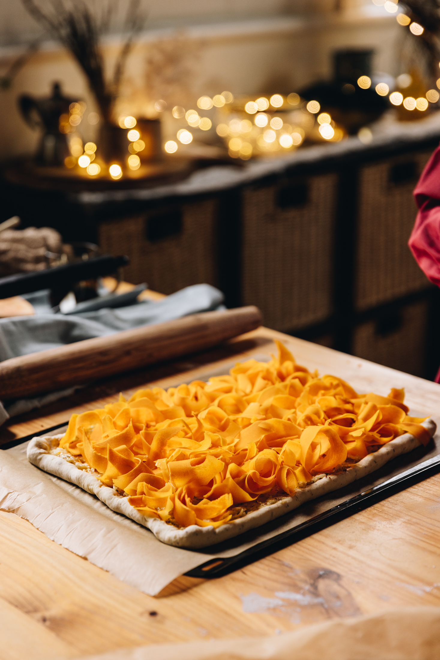 A black oven tray lined with brown baking paper sits on a wooden table. On the tray is a large pastry tart covering this whole tray with ribbons of pumpkin on top of it. In the background is blurred fairy lights.