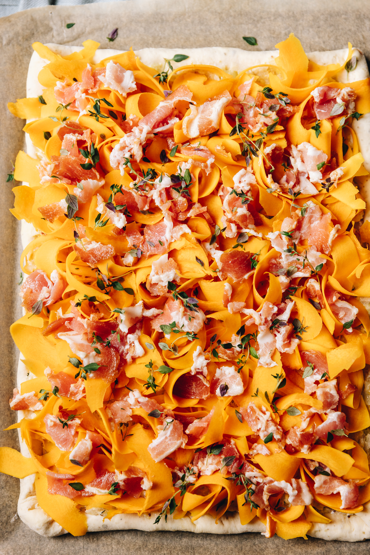 A flat lay shot shows am unbaked Creamy pumpkin tart that has ribbons of orange pumpkin, fresh thyme leaves, grated cheese and bacon on top a large piece of rolled out pastry.