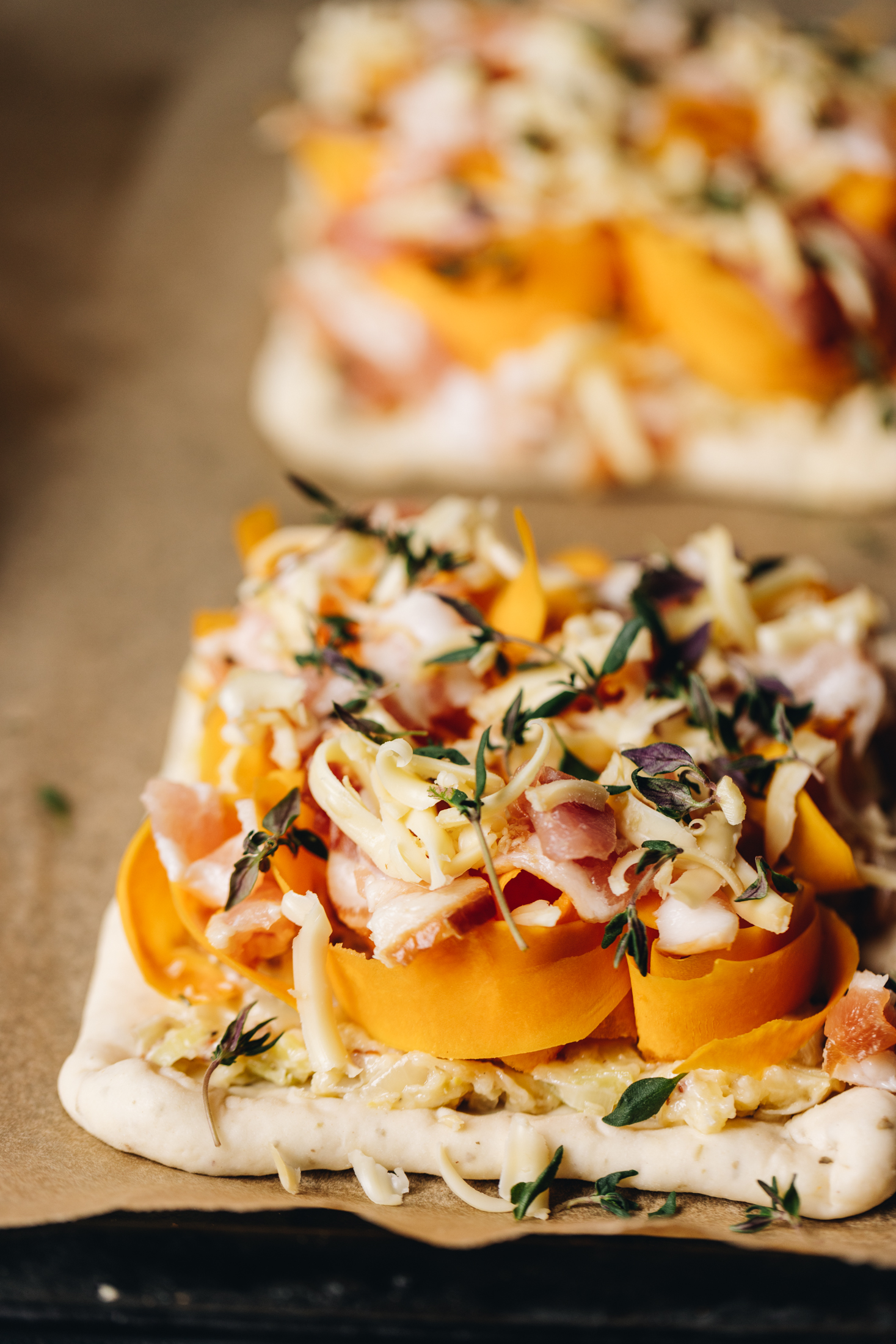 Unbaked mini creamy pumpkin tarts are on a black baking tray lined with baking paper. It has ribbons of orange pumpkin, fresh thyme leaves, grated cheese and bacon on top a large piece of rolled out pastry. Another one is blurred in the background.