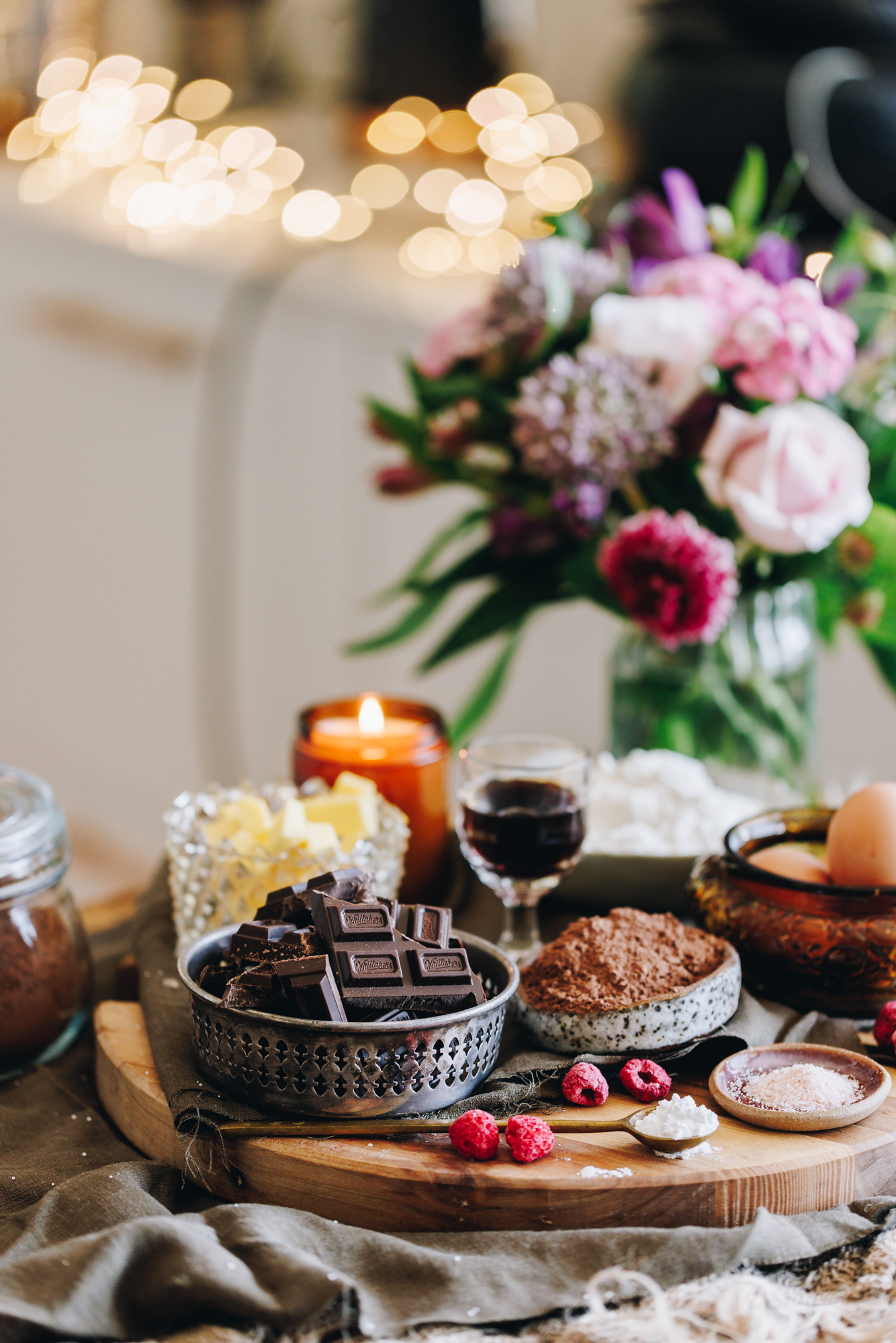 On a wooden table with natural fabrics sits a wooden board with vintage plates and crockery. There is chocolate, freeze-dried raspberries, cocoa and butter in view. An amber candle burns in the background and a bunch of flowers.