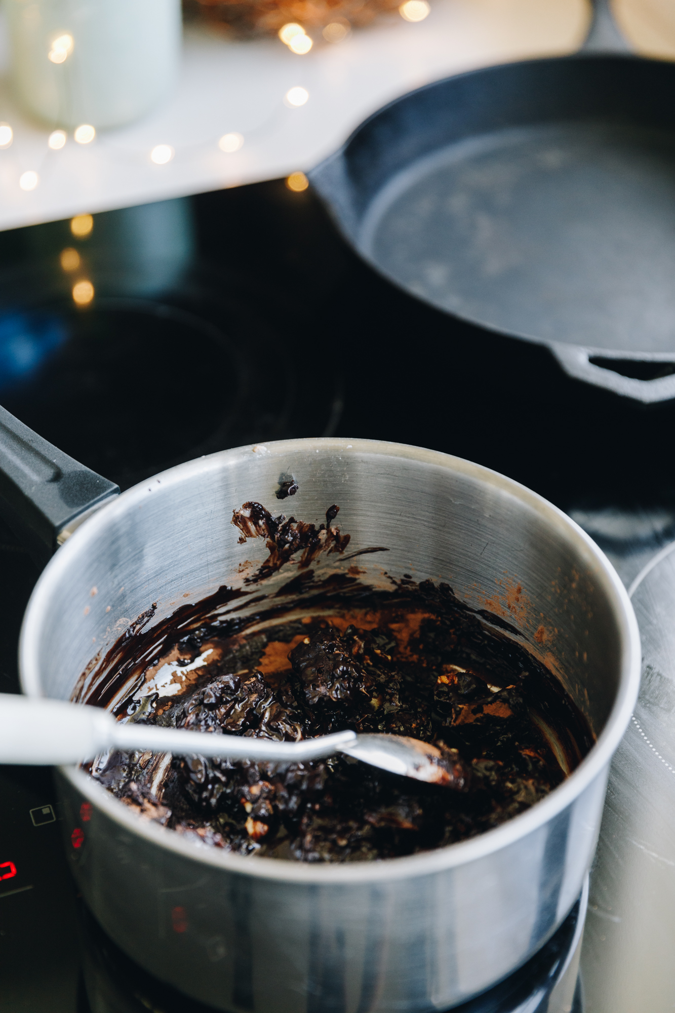 A silver pot sits on a black stove top. In the pot is a melting chocolate mixture and a spoon. A black cast iron pan and fairy lights are in the background. 