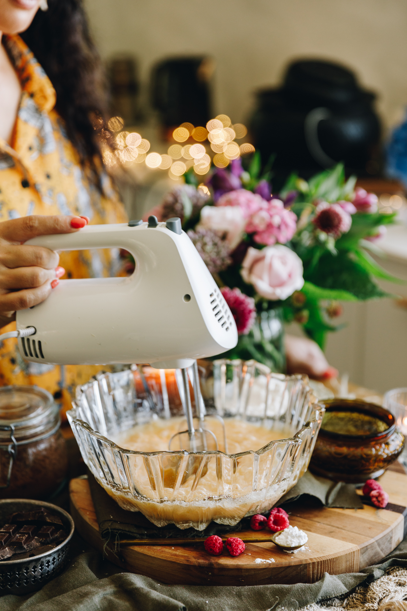 On a wooden board sits a glass vintage bowl. Inside is eggs being whipped with a white hand beater held by Naomi Toilalo. Flowers are in the background. 