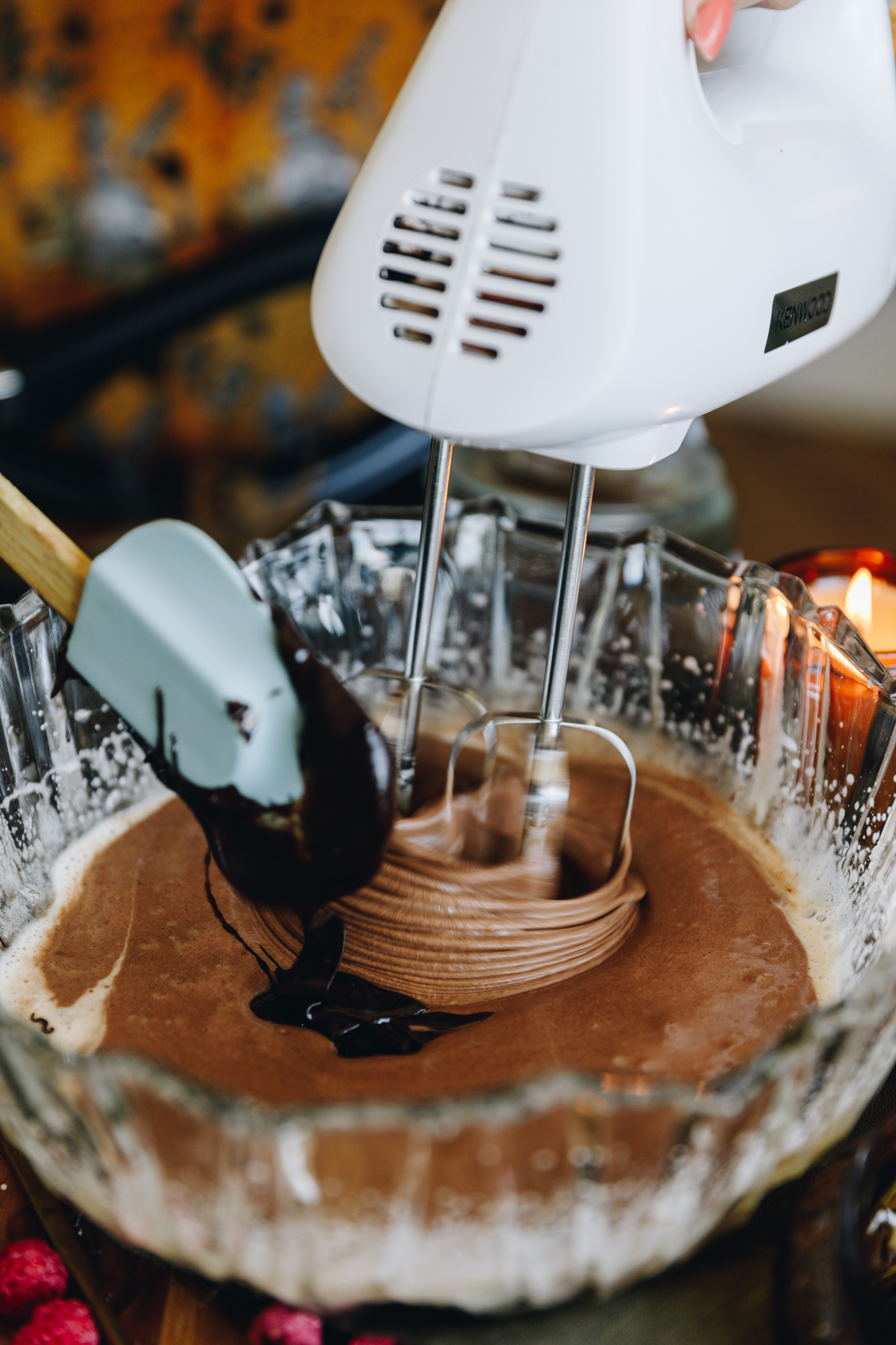 A glass vintage bowl sits on a wooden board. In the bowl is an egg mixture that is being whipped with a white hand mixer. A blue spatula is adding in a melted chocolate mixture as it is being whipped. 