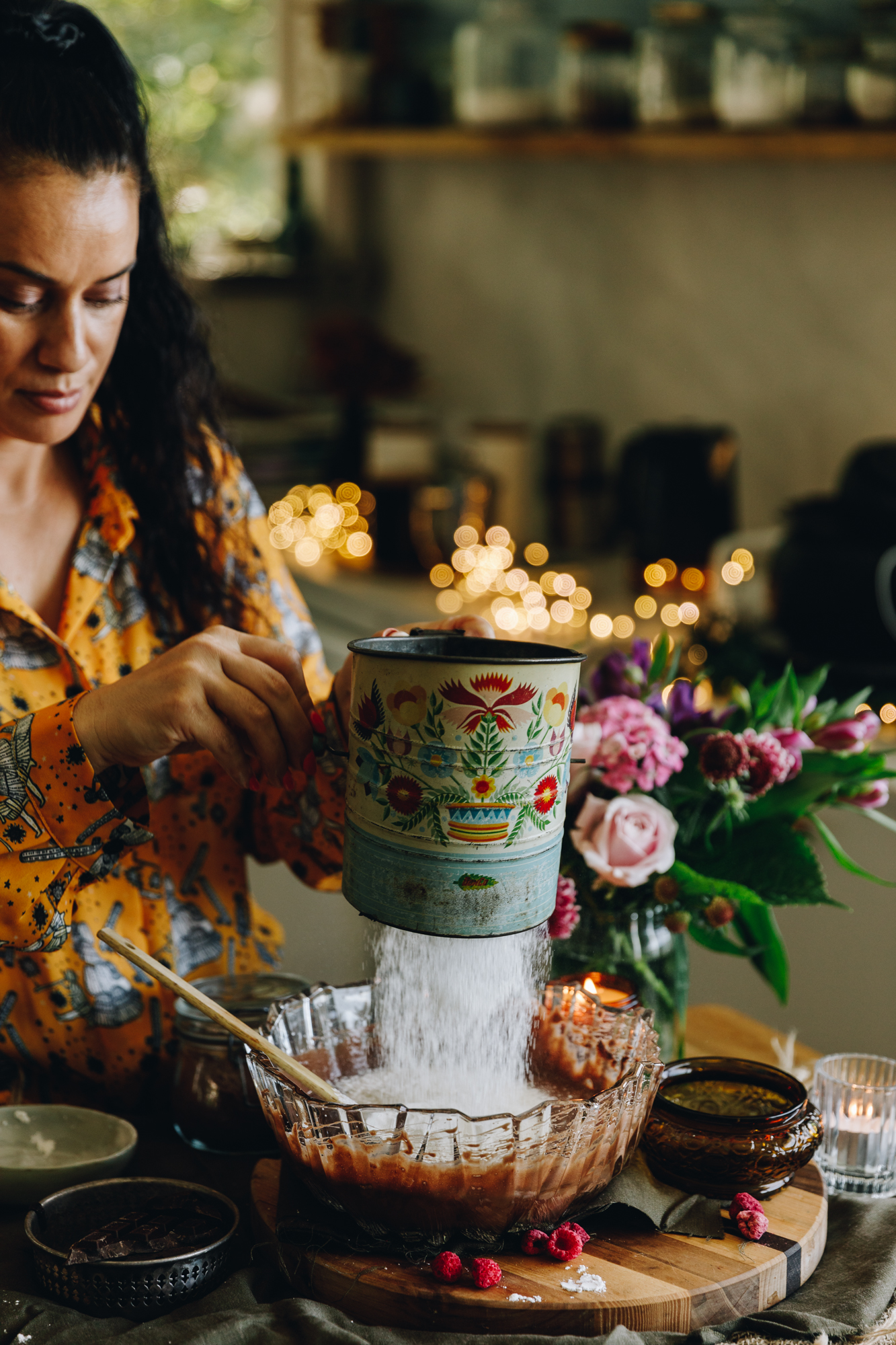A glass vintage bowl sits on a wooden board. In the bowl is whipped chocolate mixture. Naomi Toilalo is sifting flour in to the bowl with a vintage sifter. 