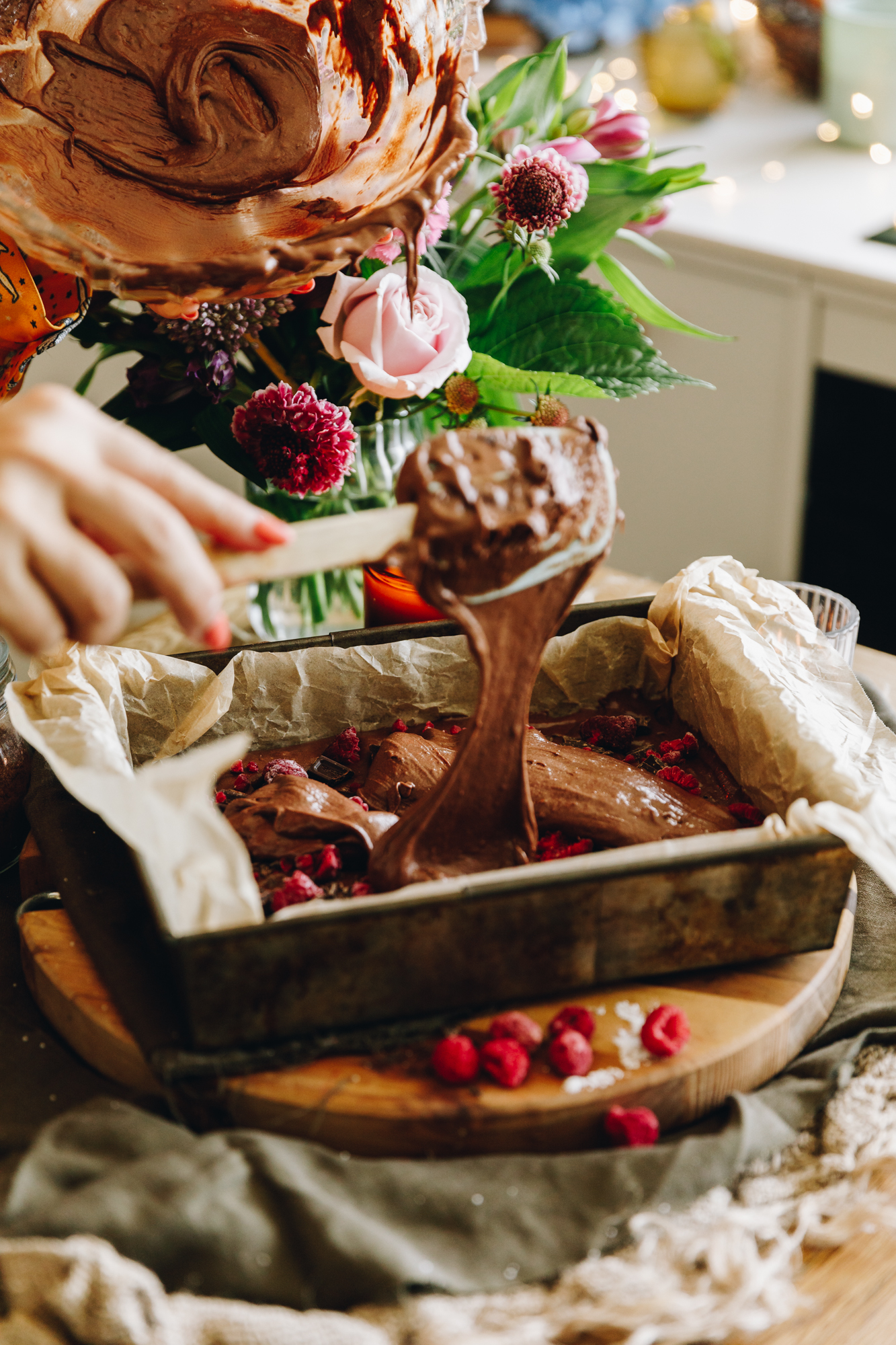 A baking tray lined with brown baking paper is on a wooden board resting on a green tablecloth. In the tin is chocolate and raspberry brownie mixture that is being layered with raspberries and chocolate chunks. Naomi is adding more mixture on top and a glass bowl with a little bit of mixture is seen. 