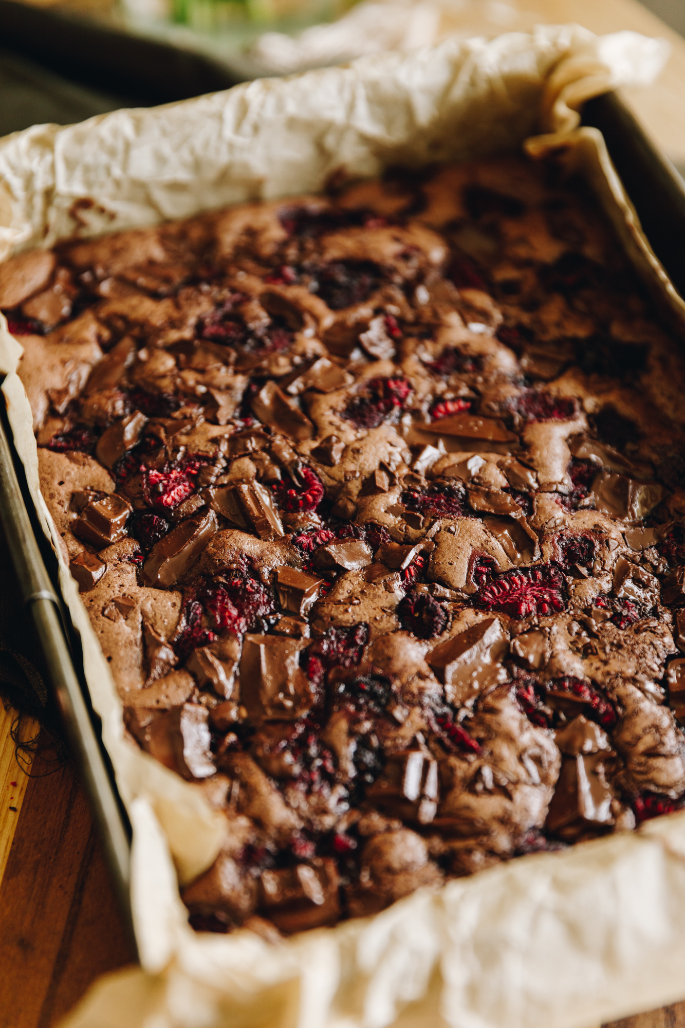 A close up shows a freshly baked chocolate and raspberry brownie that is in a baking tin lined with borne baking paper. 