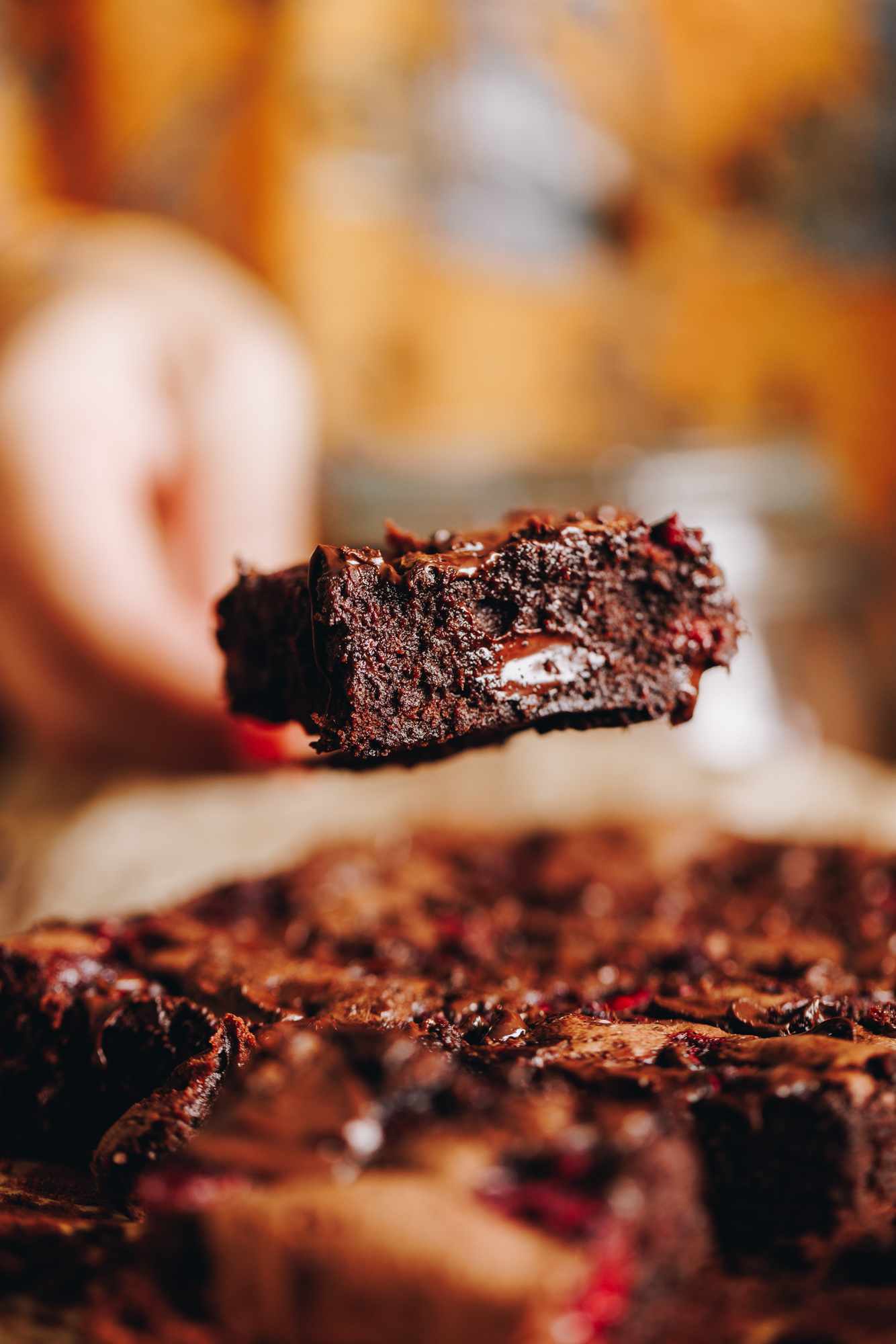 A piece of chocolate and raspberry brownie is held up in the air by a hand. It shows gooey chocolate and the moist texture of the brownie. 