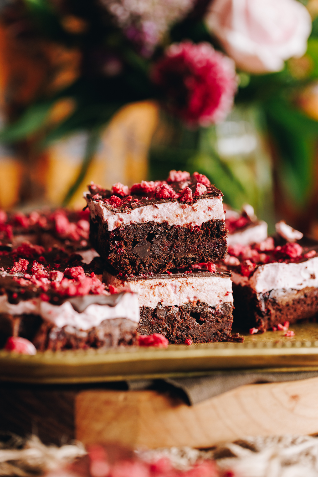On a gold tray sits cut up slices of chocolate raspberry brownie with marshmallow. Flowers are in the background.