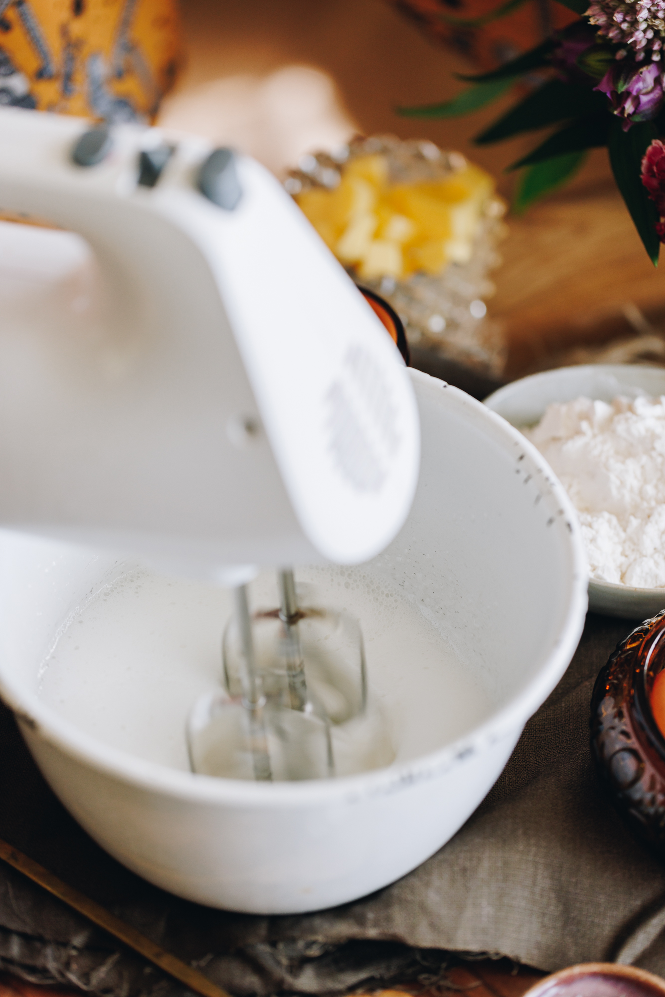 A white bowl is sitting on natural fabric. It has a meringue inside it that is being whipped by a white hand beater.