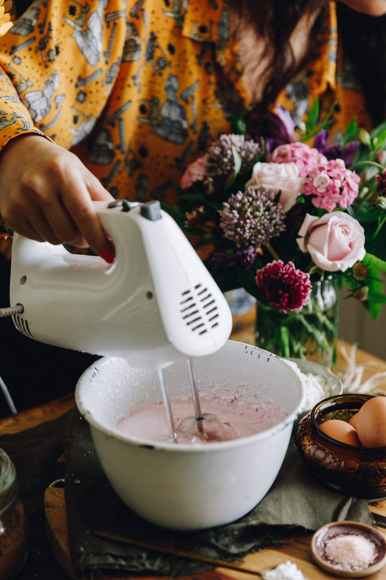 A white bowl is sitting on natural fabric. It has a raspberry meringue inside it that is being whipped by a white hand beater. A bunch of flowers sit behind the bowl.
