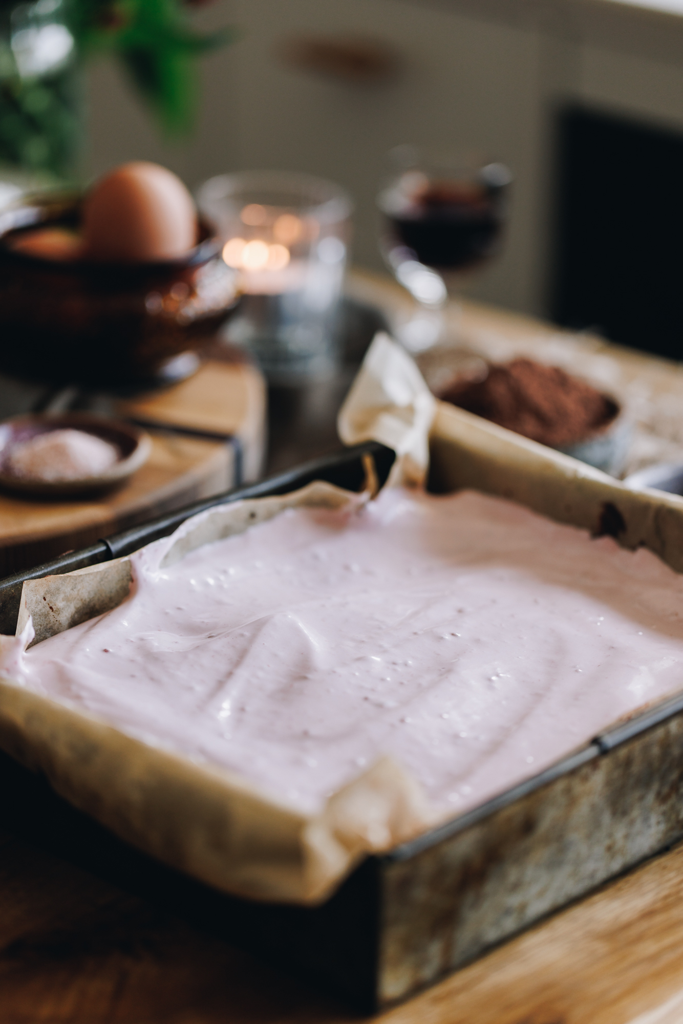 A baking tin lined with brown baking paper sits on a wooden table. It has fresh raspberry marshmallow that has been spread on top of the slice.