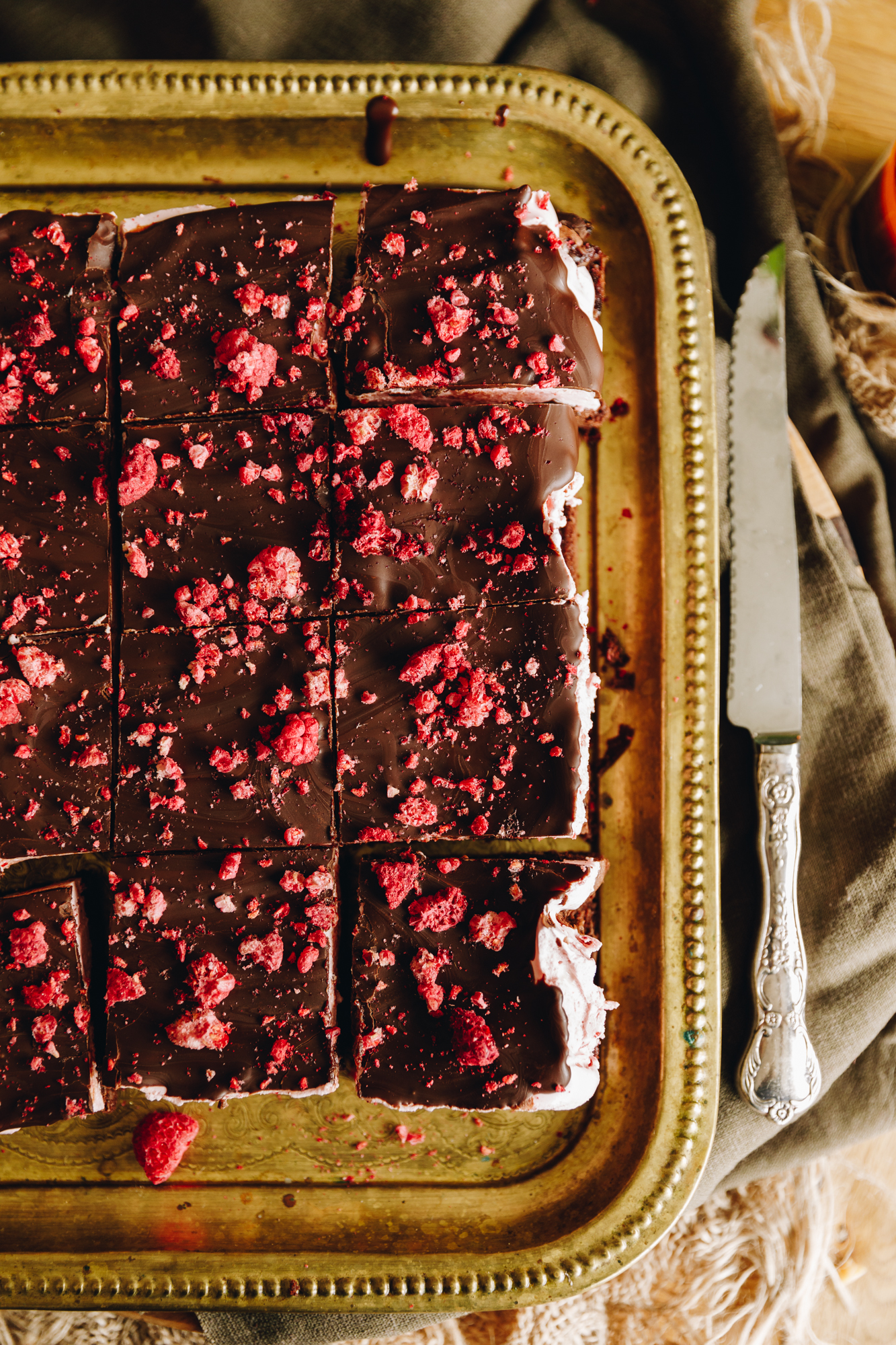 A top shot shows a fresh decorated chocolate raspberry brownie with marshmallow that has been decorated with dark chocolate and freeze-dried raspberries. It has been sliced in to pieces. The gold tray is on top of natural fabrics.