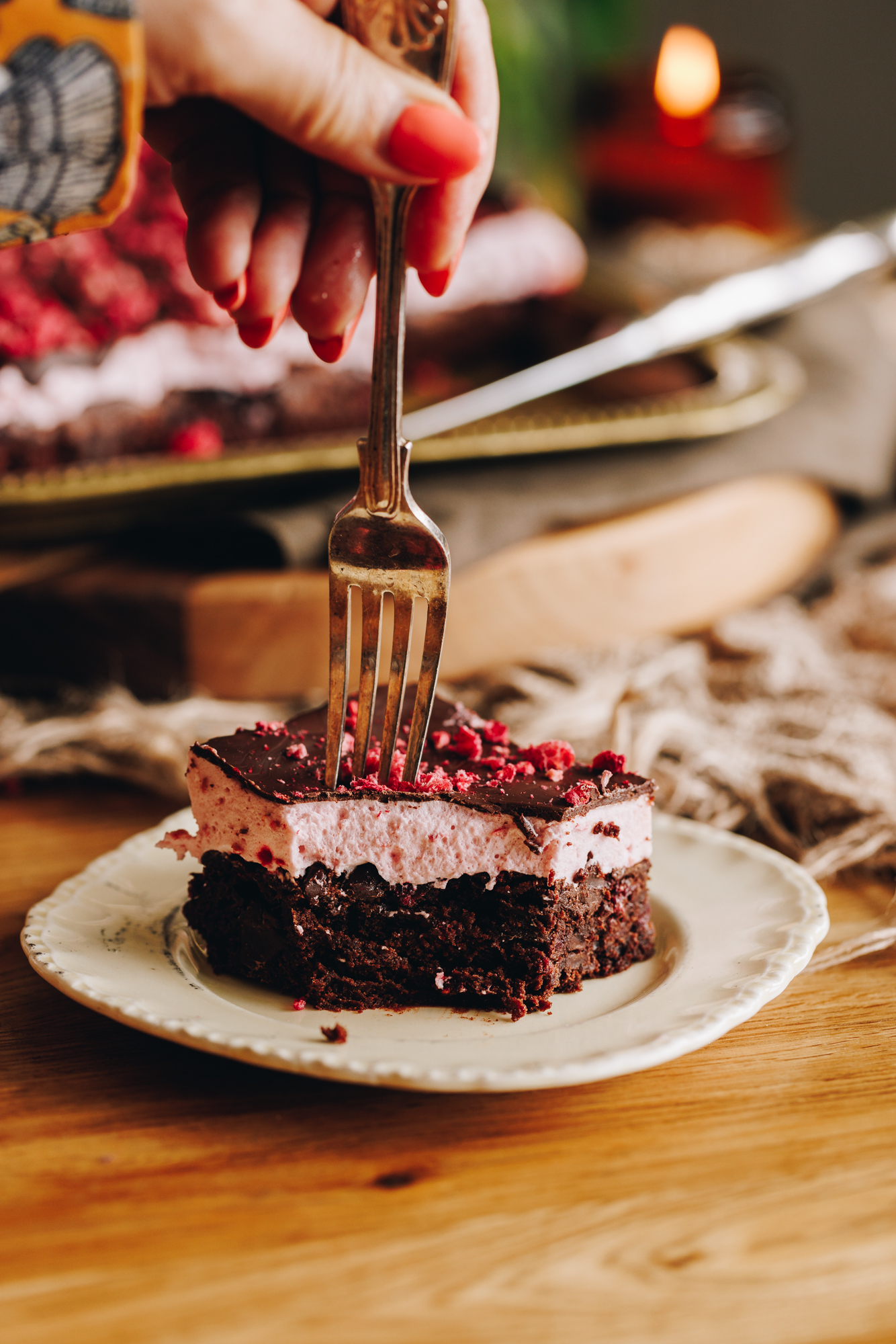A slice of chocolate raspberry brownie with marshmallow that has been decorated with dark chocolate and freeze-dried raspberries sits on a cream vintage plate. A fork is going through it. It is on a wooden table.