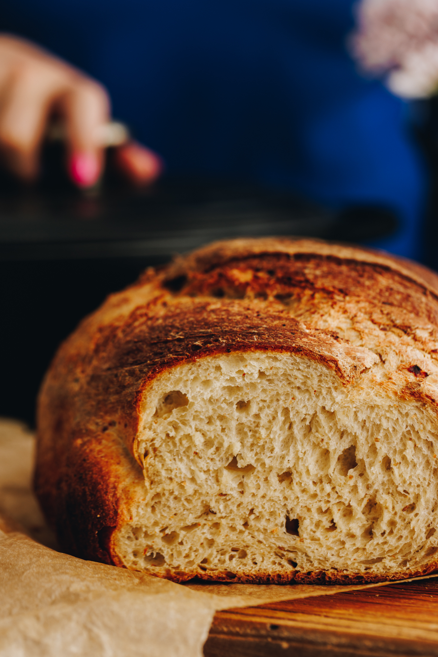 A freshly baked cottage cheese cumin loaf is sitting on a wooden board with brown baking paper on it. A hand in the background is holding the handle of a black cast iron pot. 