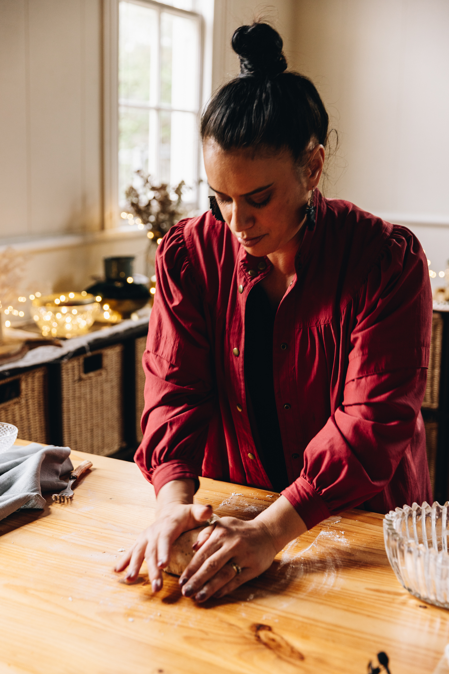 Naomi Toilalo is standing at a wooden table and is kneading dough. There is a tablecloth next to her and fairy lights in the background.