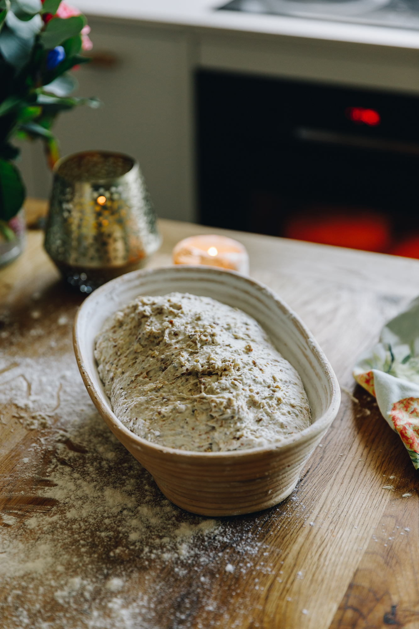 On a floured wooden table is a sourdough banneton that has an unbaked dough in it. Behind it a candle and flowers in a vase with the oven in the background. 