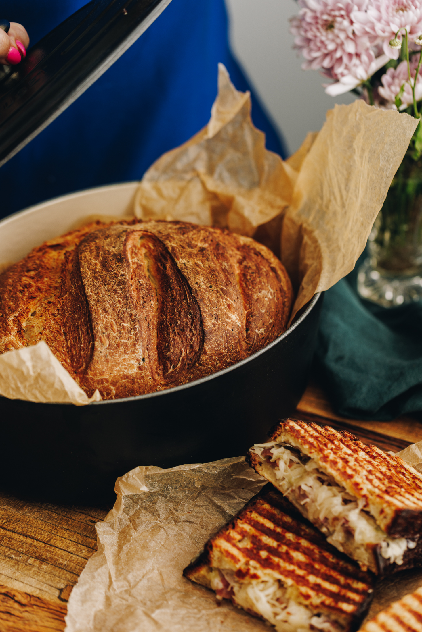 On a wooden table is a black cast iron pot with freshly baked cottage cheese bread recipe. Baking paper lines the pot and a maple bacon cheese toastie is seen in the corner of the shot. 