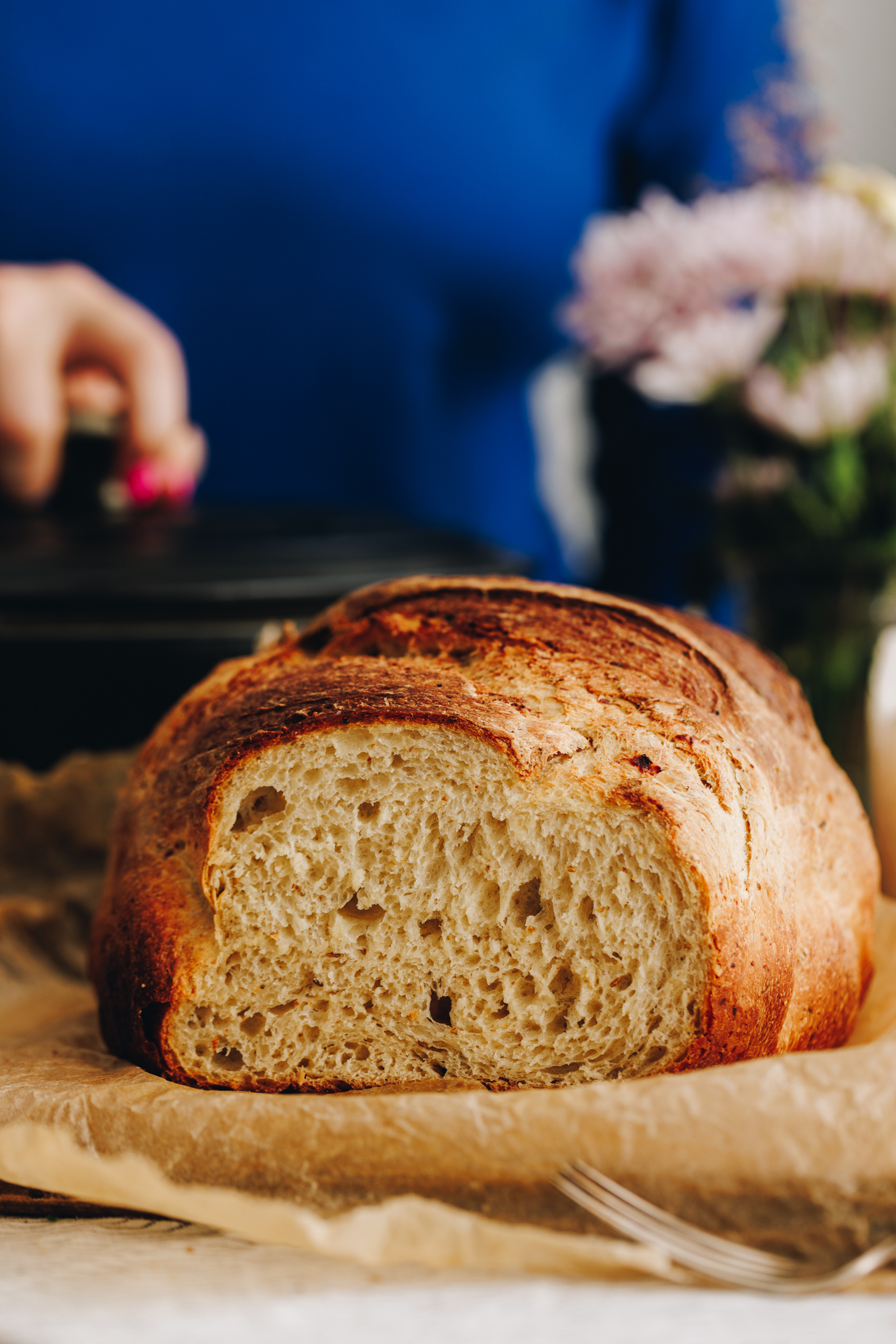 A freshly baked cottage cheese cumin loaf is sitting on a wooden board with brown baking paper on it. A hand in the background is holding the handle of a black cast iron pot.