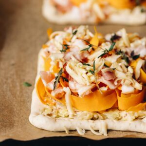 An unbaked mini creamy pumpkin tart is sitting on brown baking paper. It has ribbons of pumpkin with pieces of bacon, grated cheese and fresh thyme on it.