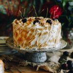 A whole Gluten-free Almond Sponge is on a ceramic cake stand. It is topped with swirls of toasted meringue and fresh cherries.