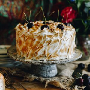 A whole Gluten-free Almond Sponge is on a ceramic cake stand. It is topped with swirls of toasted meringue and fresh cherries.